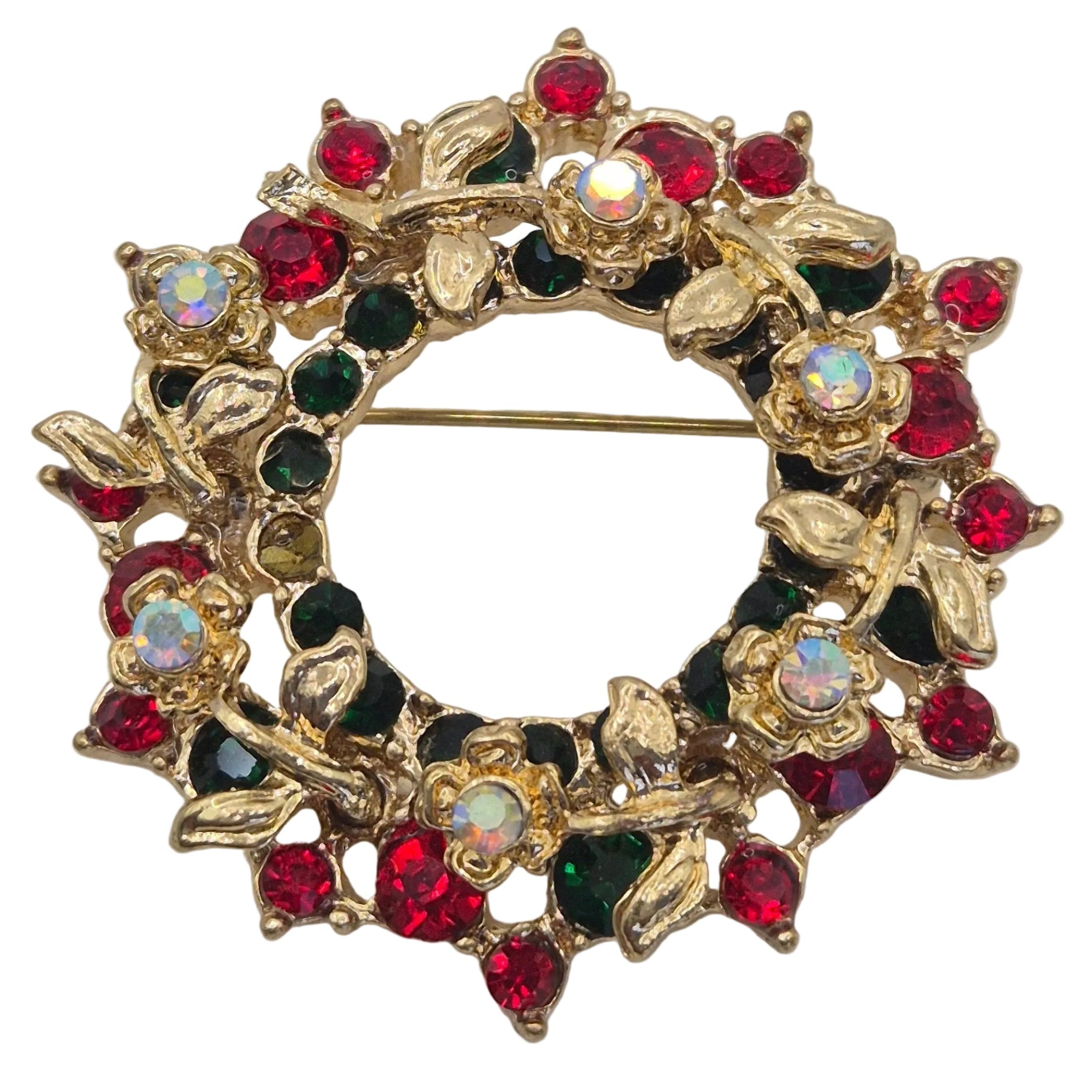 A gold-tone floral wreath brooch embellished with red, dark green, and clear rhinestones, displayed on a neutral light gray background. Close up view.