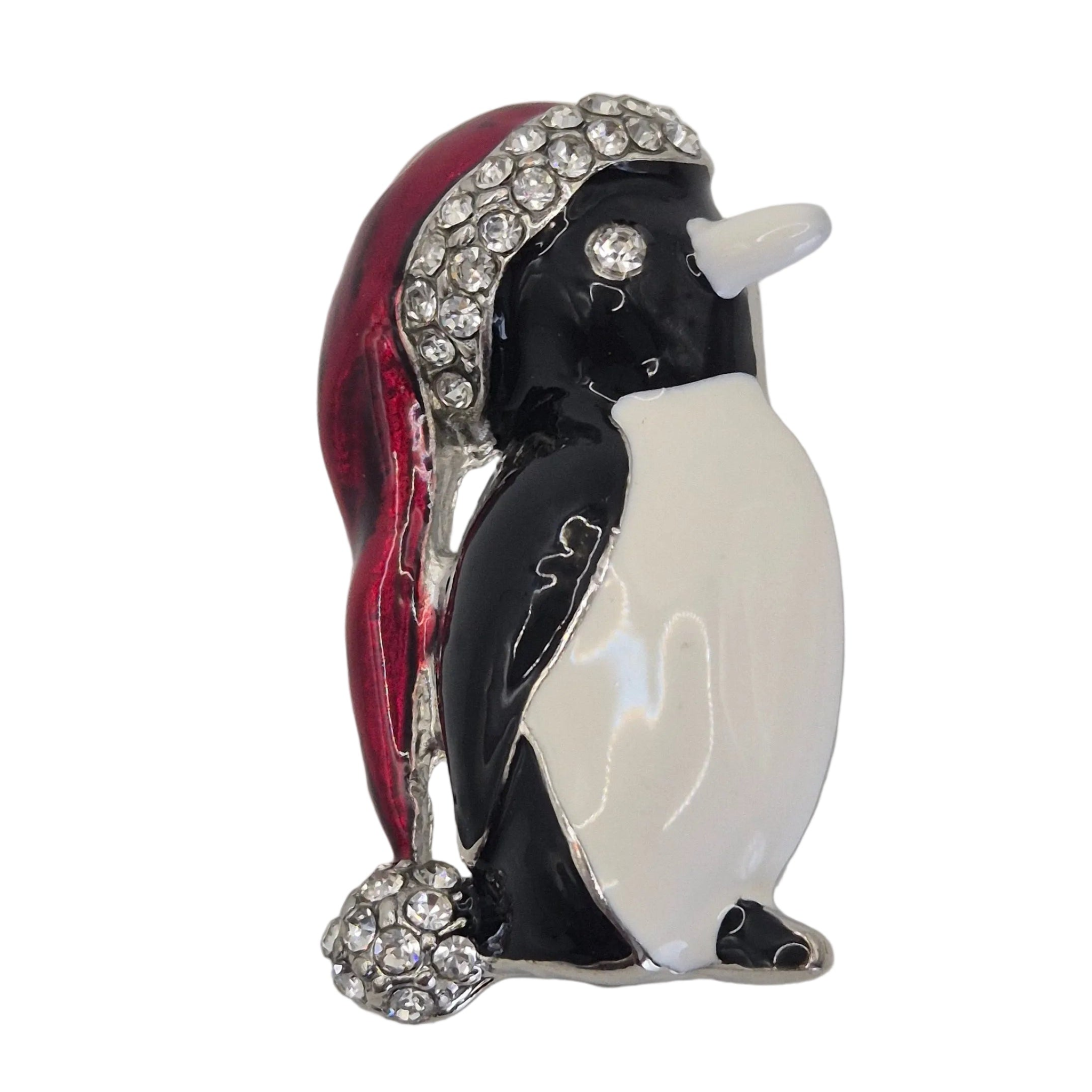 A penguin-shaped brooch featuring black and white enamel, a red Santa hat with rhinestone trim, and a small rhinestone ball, shown on a plain white background. Close up shot.
