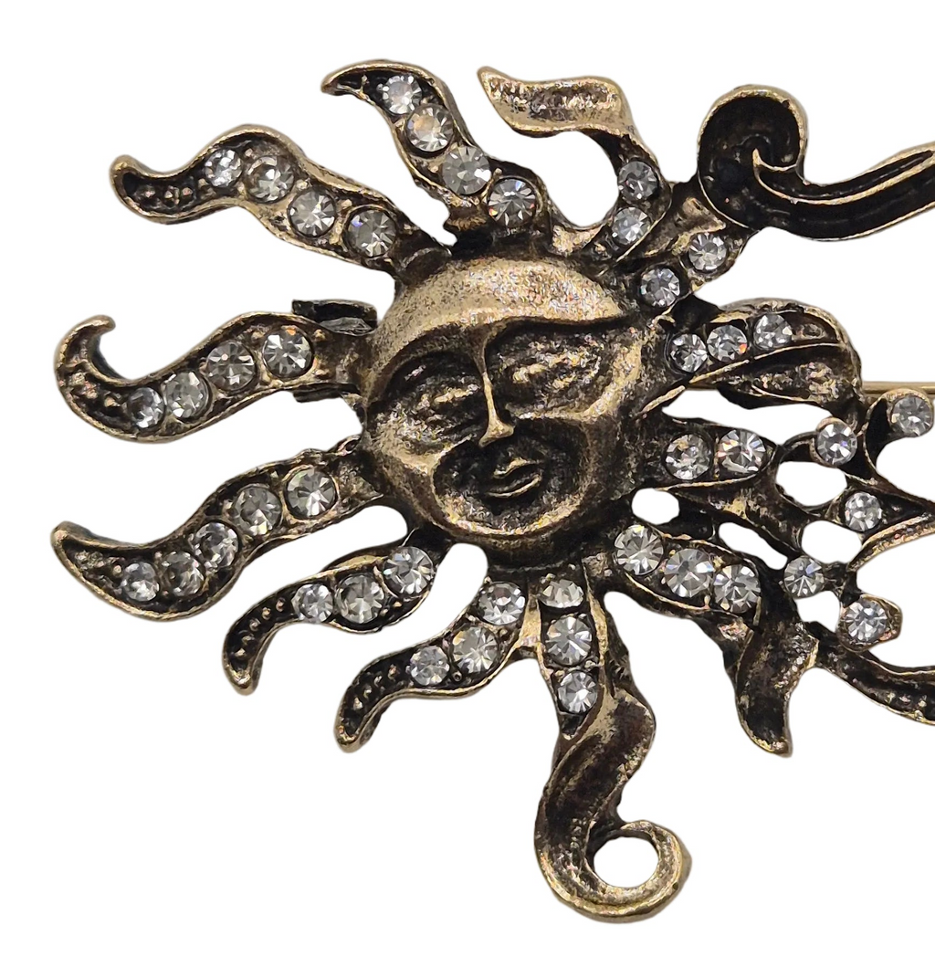 An antique sun and moon brooch showing sun with a smiling face and rays studded with clear rhinestones, shown on a plain white background.