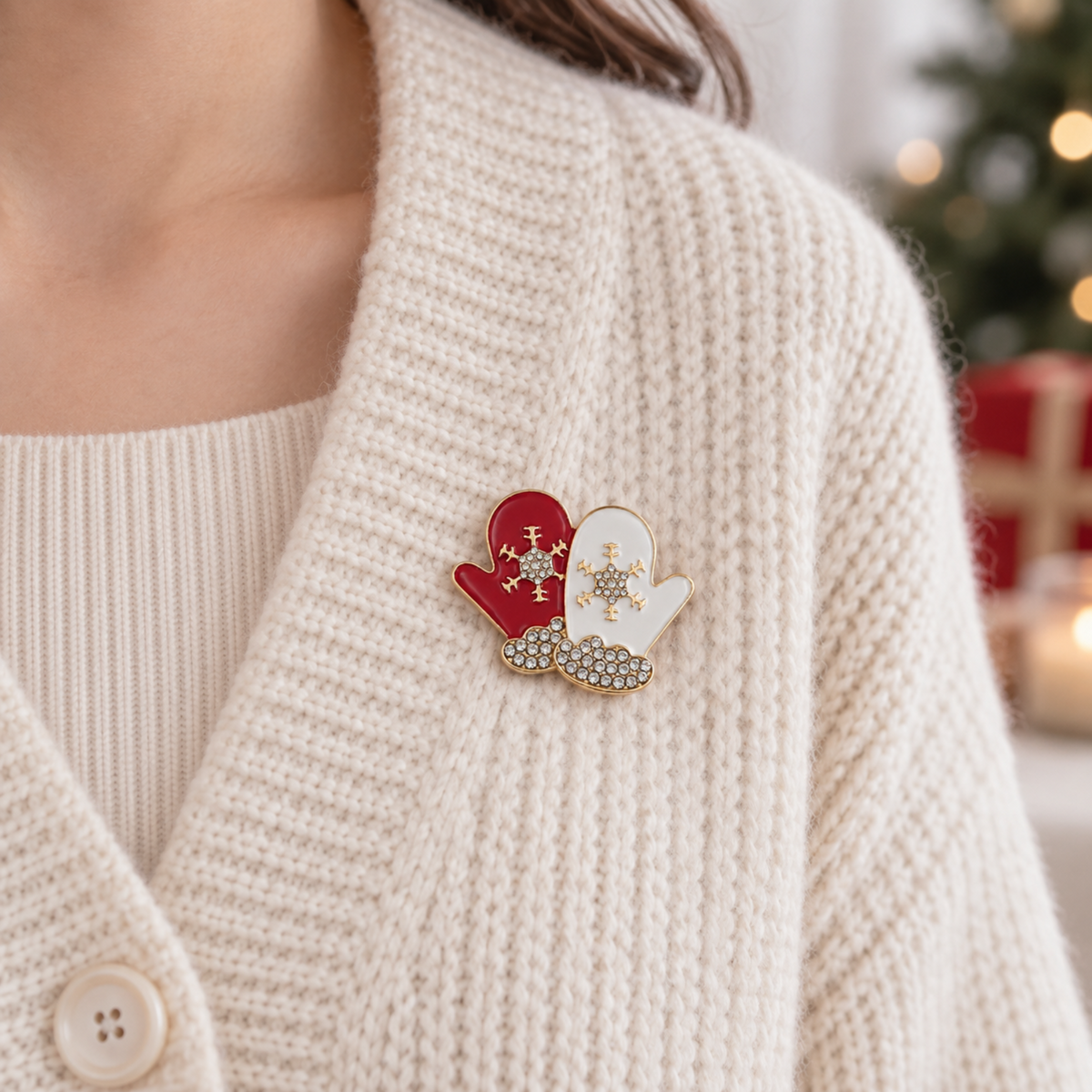 A close-up of a person wearing a cream knit cardigan and a small enamel pin shaped like two winter mittens, one red and one white, with gold-tone outlines and rhinestone details.