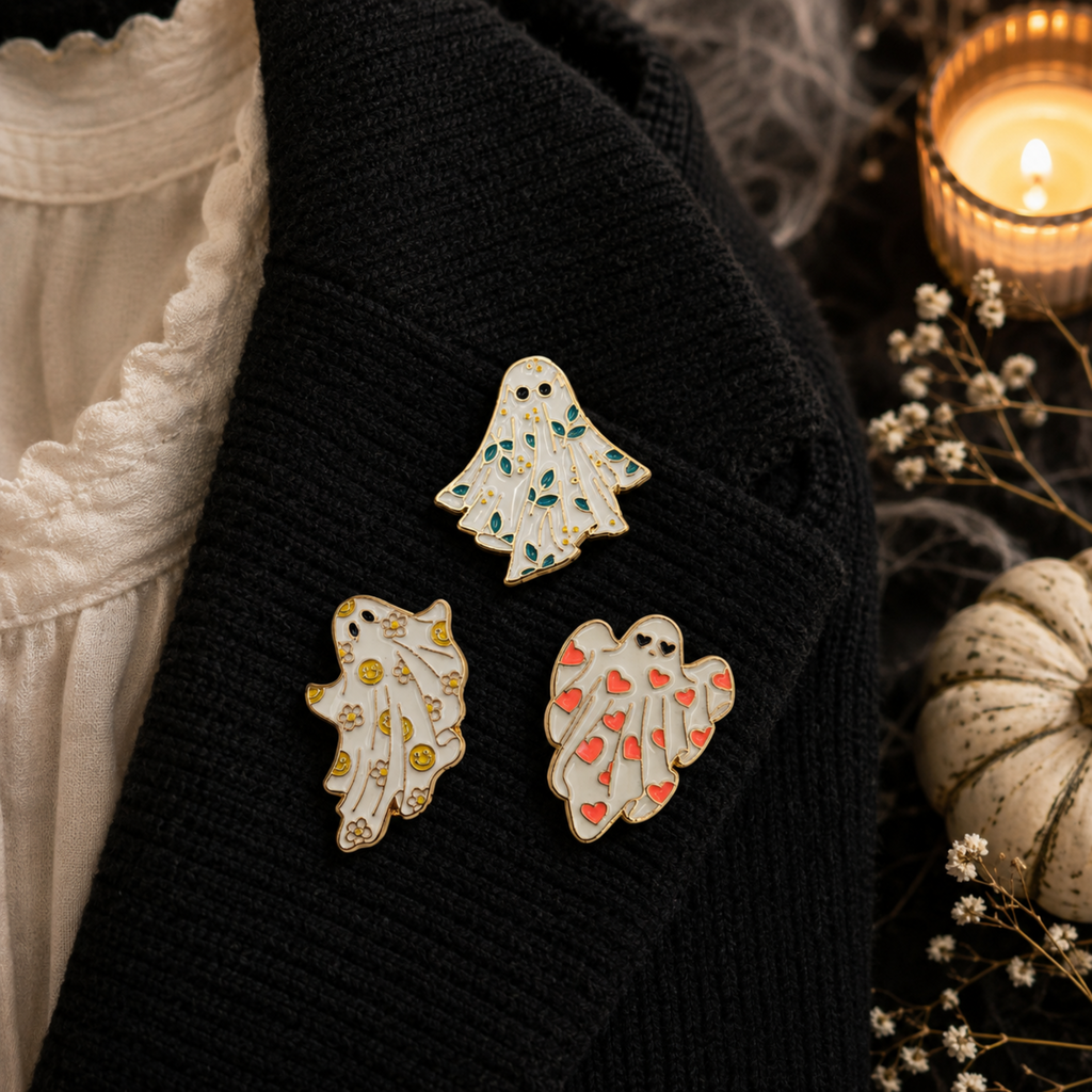 Three ghost-shaped enamel pins with Halloween-themed designs are displayed on a black knit sweater. The background features a white lace-trimmed blouse, a lit candle, and a small decorative pumpkin, creating a cozy autumn atmosphere.