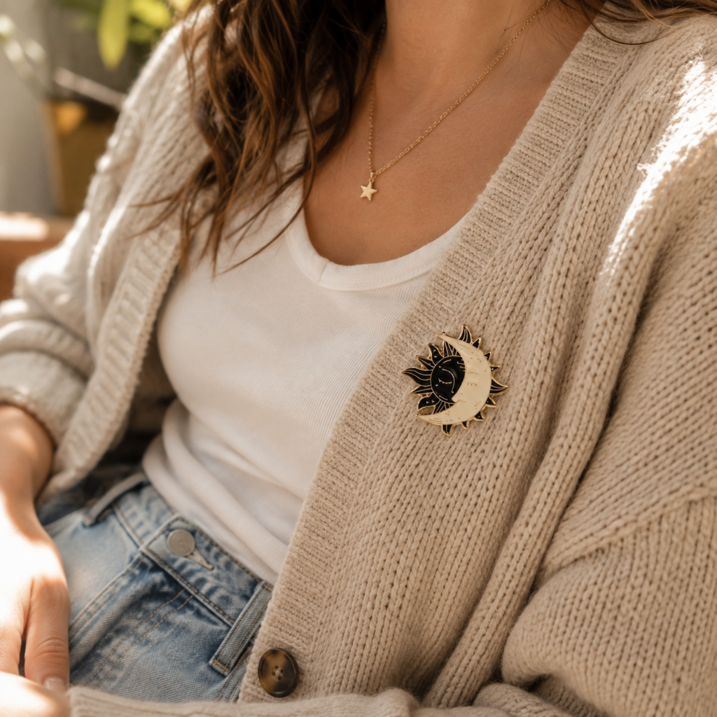 A close-up lifestyle shot of a person wearing a beige knit cardigan over a white t-shirt and blue jeans, featuring a black and gold crescent moon and sun brooch on the cardigan.