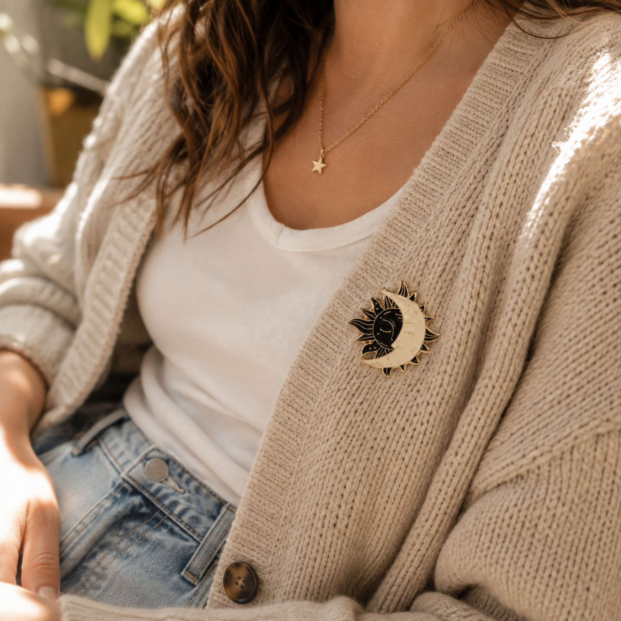 A close-up lifestyle shot of a person wearing a beige knit cardigan over a white t-shirt and blue jeans, featuring a black and gold crescent moon and sun brooch on the cardigan.