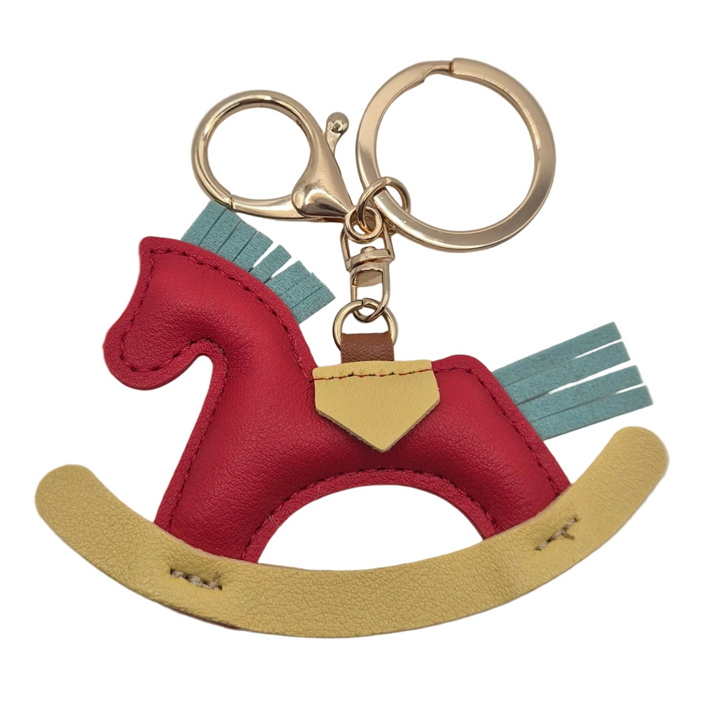 A close up shot of a small rocking horse keychain charm made from red, light blue, and yellow faux leather, attached to gold-tone metal rings and a lobster clasp, photographed on a plain white background.