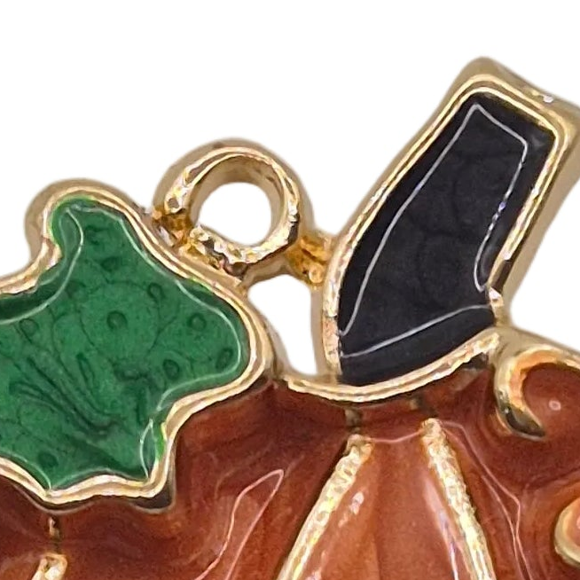 Close-up of a gold-tone enamel pumpkin brooch charm featuring a green leaf, a black leaf, and a terracotta-colored body with gold trim on a white background.