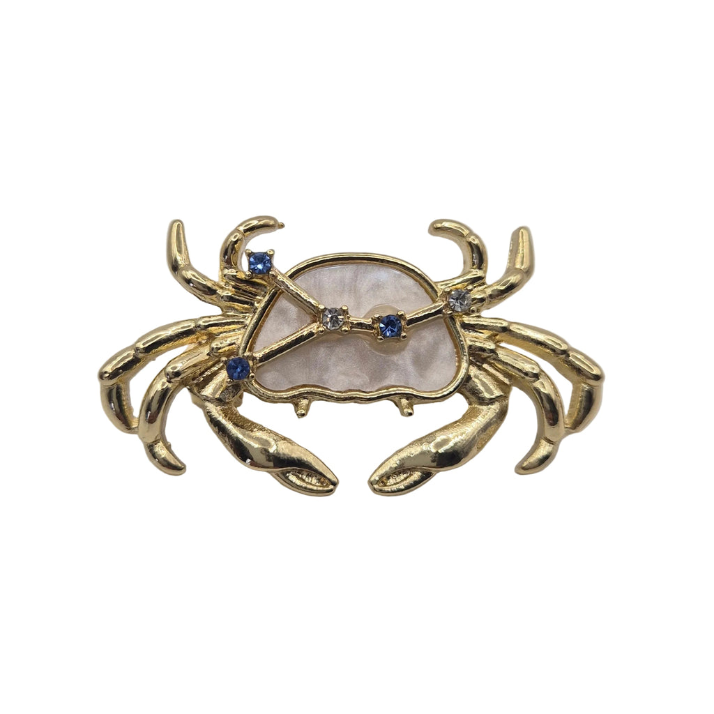 A gold crab-shaped brooch featuring a mother-of-pearl shell inlay and small blue gemstone accents, displayed on a plain white background.