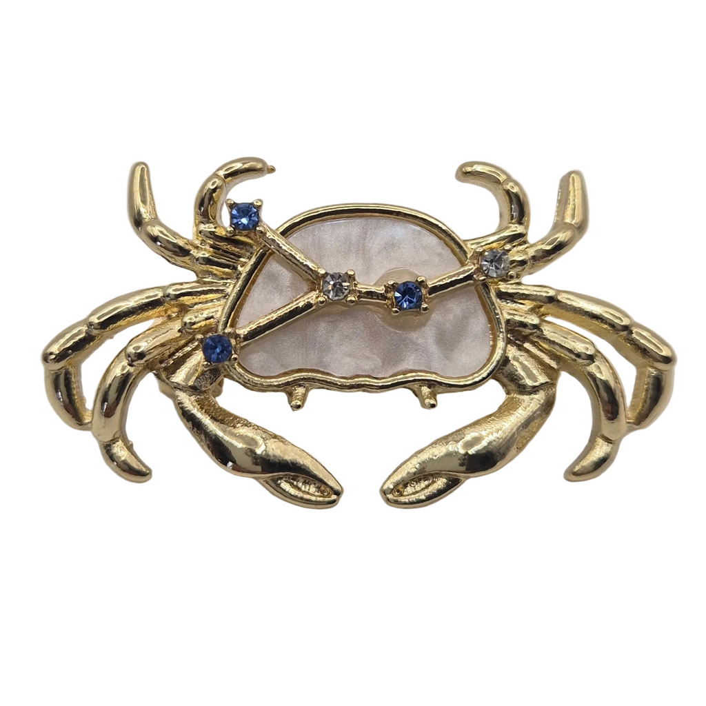 A close up shot of a gold crab-shaped brooch featuring a mother-of-pearl shell inlay and small blue gemstone accents, displayed on a plain white background.