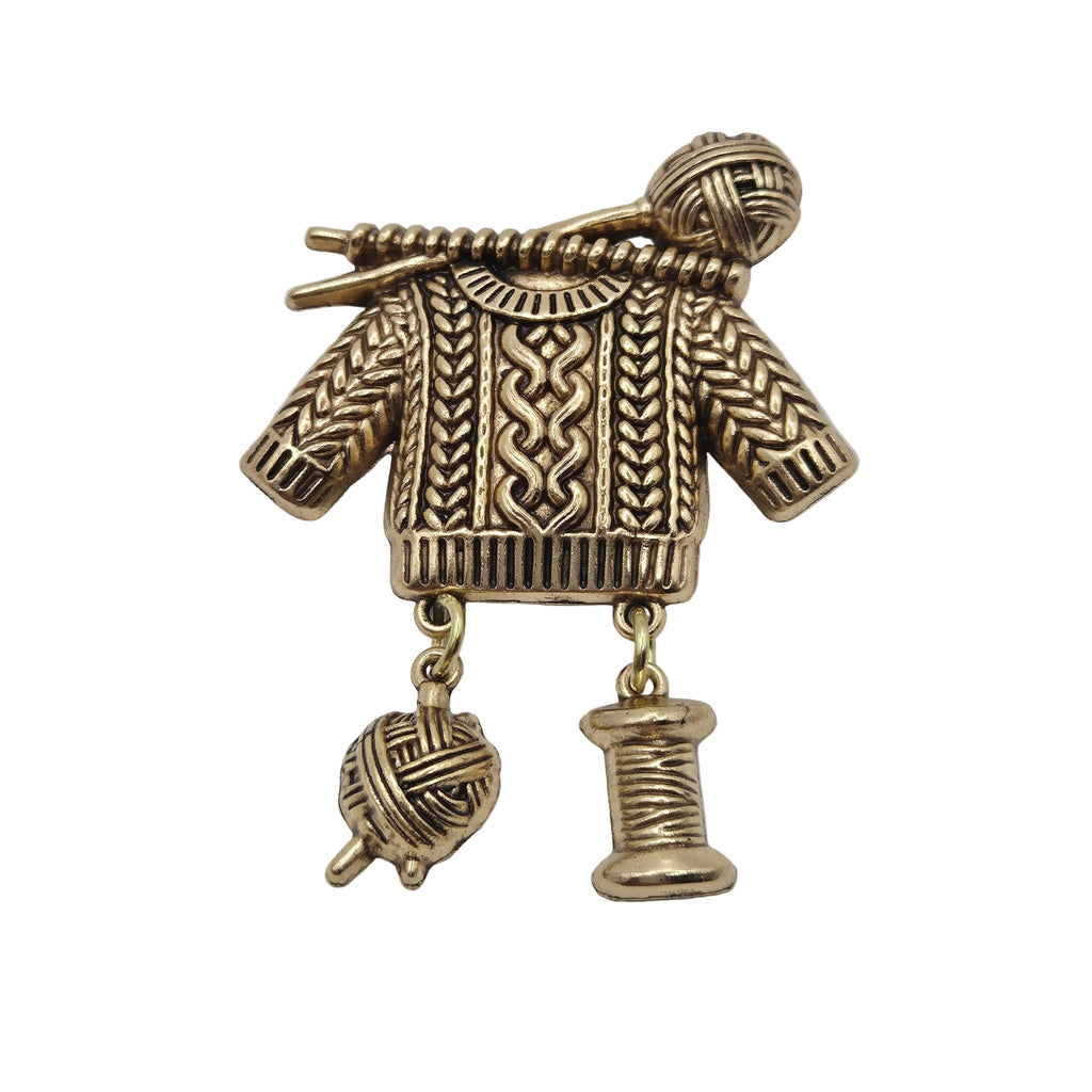 An antique gold tone metal pendant shaped like a knitted sweater with knitting needles and yarn balls, shown on a plain white background.