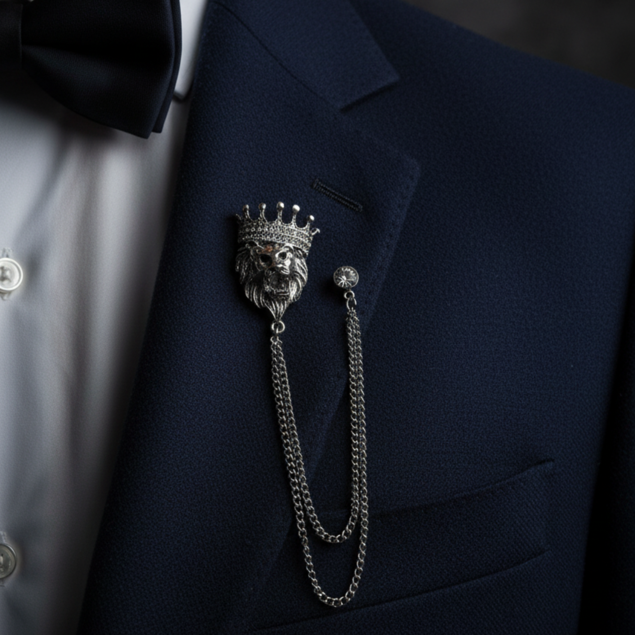 A silver lion head with a crown and chain brooch pinned to a navy blue suit jacket over a white dress shirt and black bow tie.