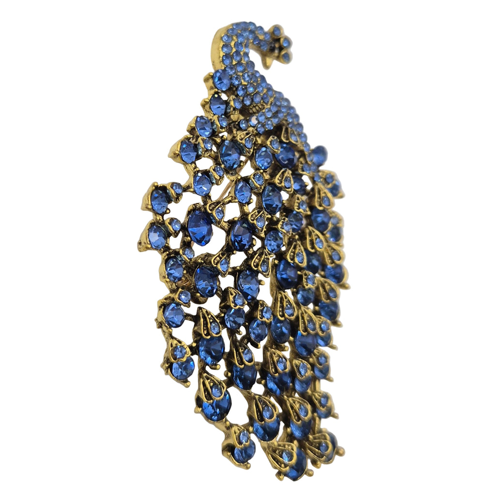 A peacock-shaped brooch featuring blue rhinestones set in gold-tone metal, photographed on a plain white background.