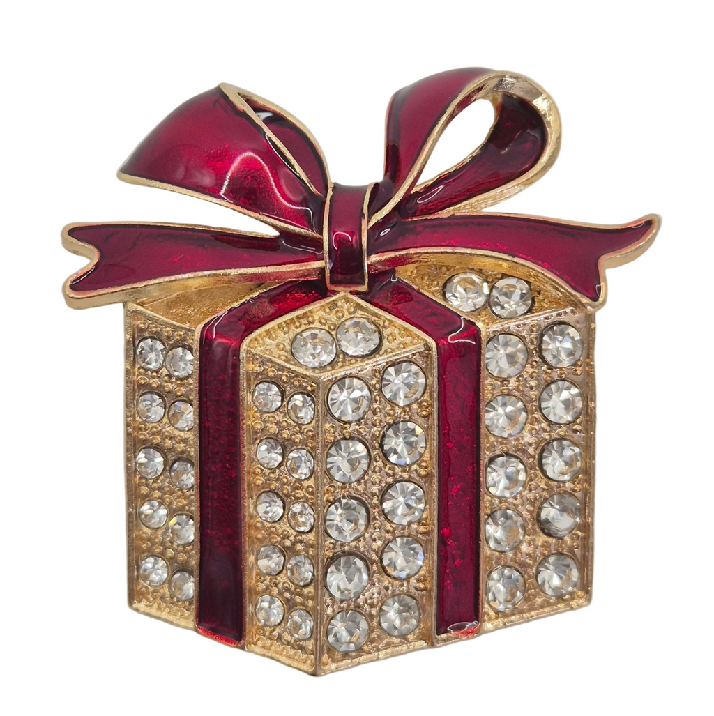 A gold-tone gift box brooch embellished with clear rhinestones and accented with a glossy dark red enamel ribbon and bow, set against a plain light white background. Close up view. 