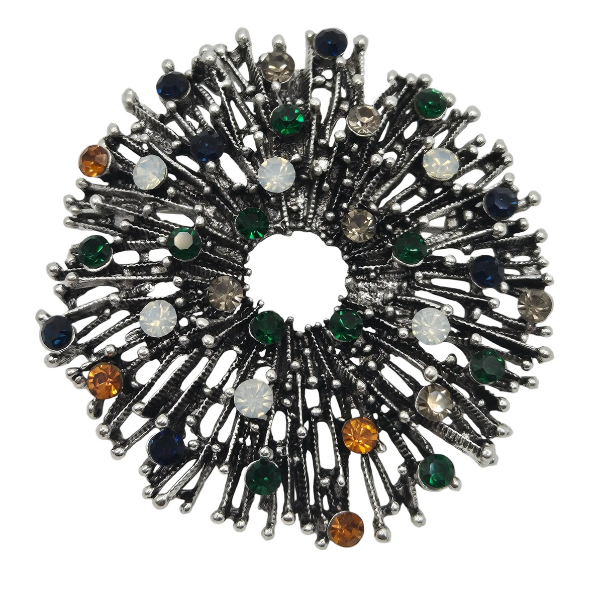 A circular brooch featuring a dark silver-toned metal firework radiating textured spikes and multiple faceted gemstones in green, blue, white, amber, and clear, set against a plain white background.