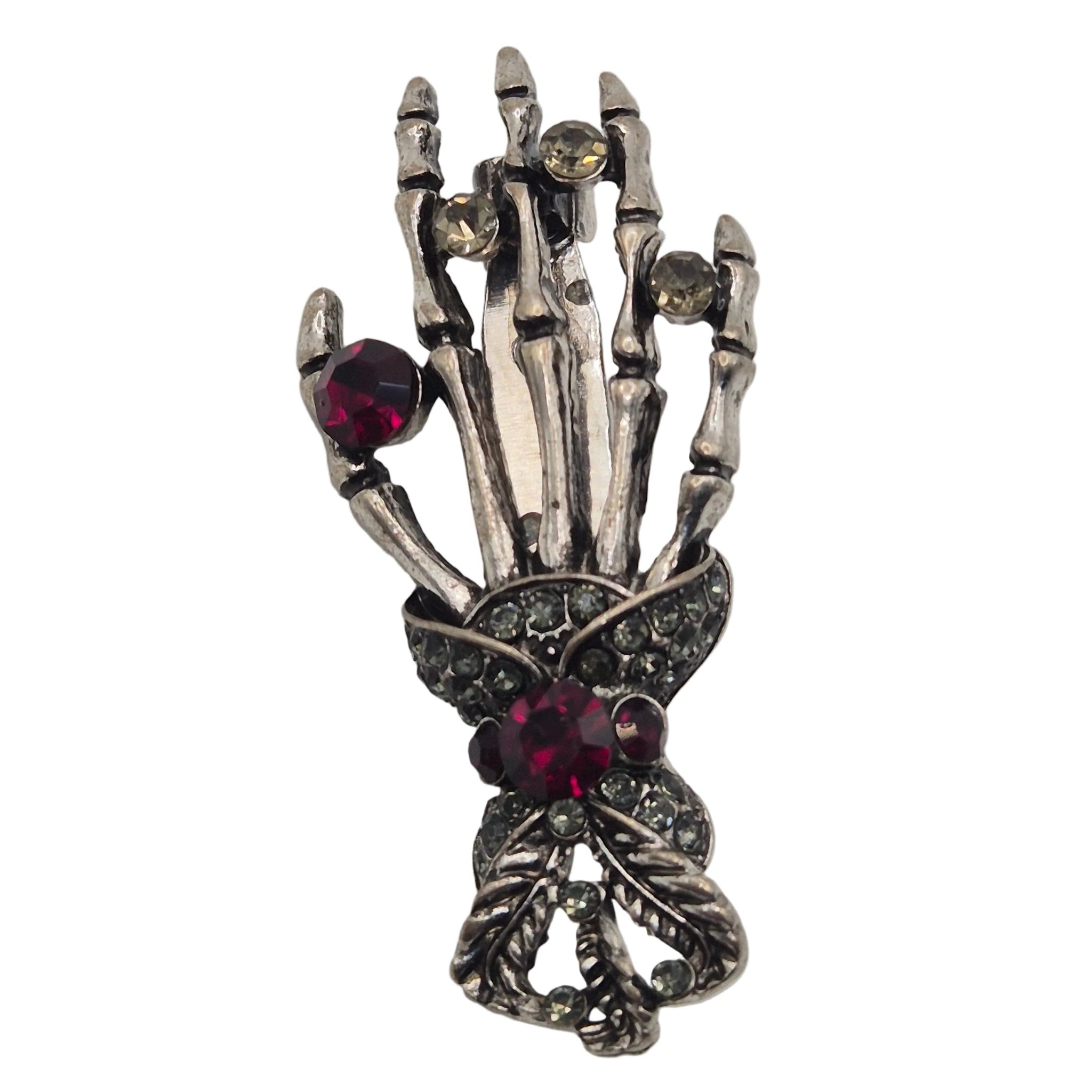 A gothic skeleton hand brooch in silver-toned metal with dark oxidized detailing, featuring red gemstone accents and small clear rhinestones, shown on a plain white background.
