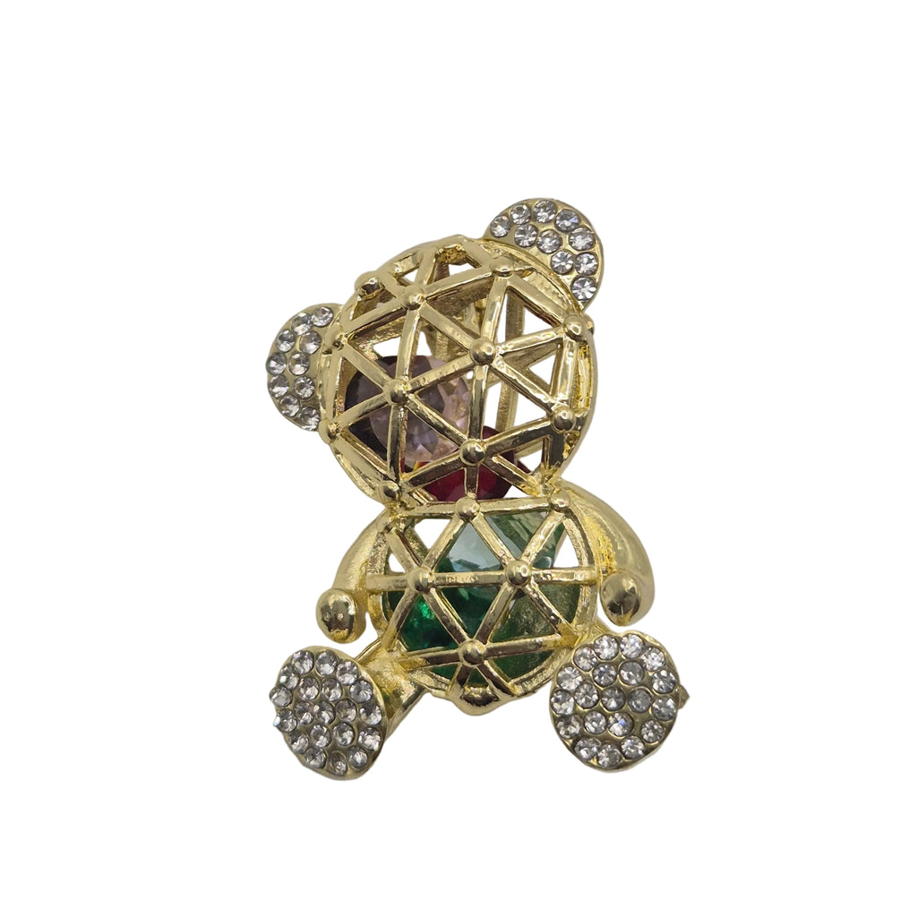 A gold-toned teddy bear brooch embellished with clear pavé crystals on its ears, feet, and chest, featuring a central faceted gemstone with green and pink inlay, set against a plain white background.