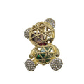A gold-toned teddy bear brooch embellished with clear pavé crystals on its ears, feet, and chest, featuring a central faceted gemstone with green and pink inlay, set against a plain white background.