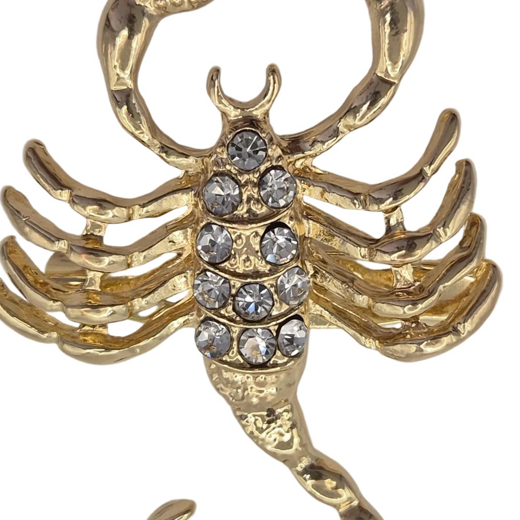 A gold-toned scorpion-shaped brooch or pendant embellished with clear rhinestones on its abdomen, shown against a plain white background.
