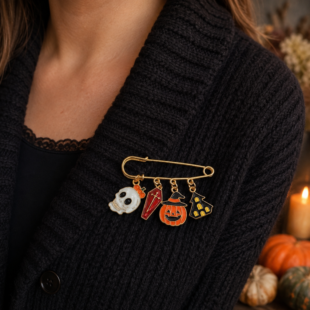A gold safety-pin style brooch with four Halloween-themed charms worn on a person's black cardigan. The charms include a white skull, a red chili pepper, an orange jack-o'-lantern, and a yellow witch hat with black dots.