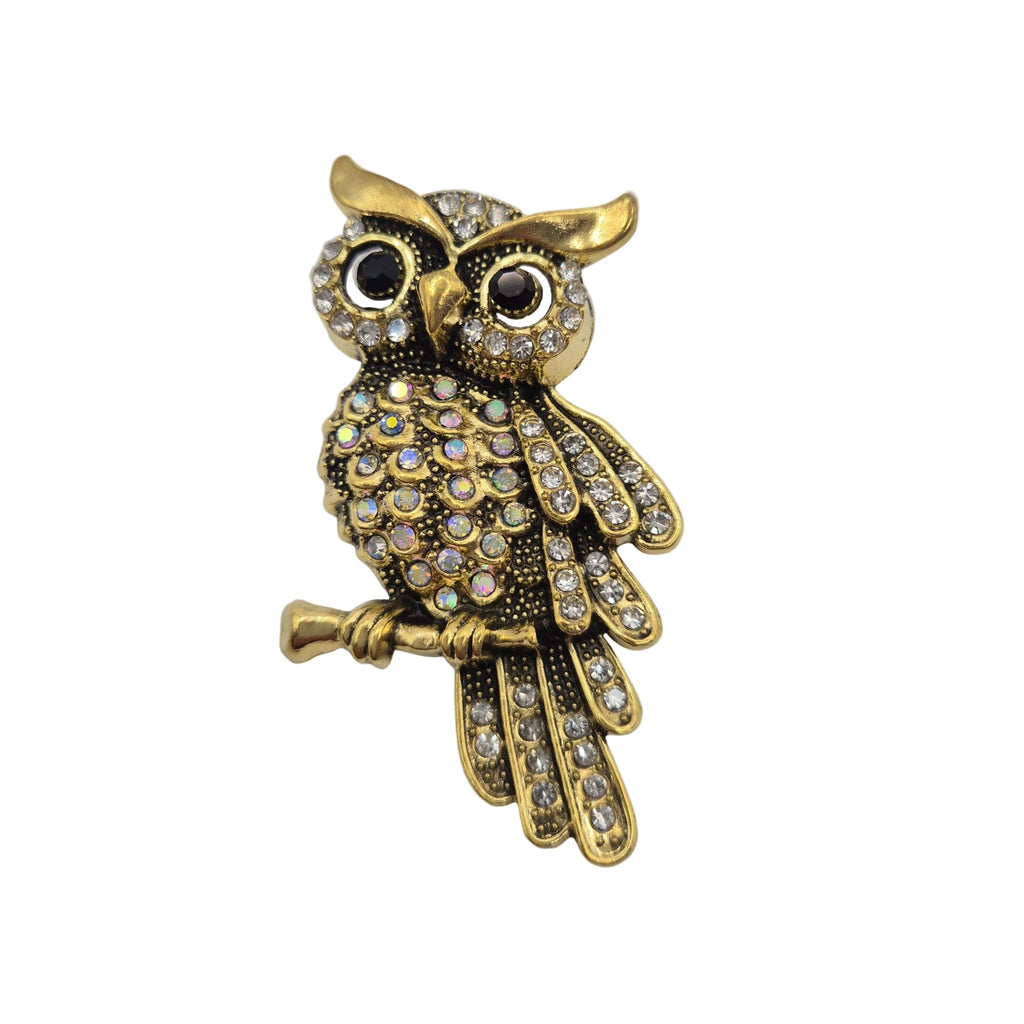 Gold owl brooch embellished with clear rhinestones, resting on a small branch, set against a plain white background.