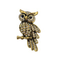 Gold owl brooch embellished with clear rhinestones, resting on a small branch, set against a plain white background.