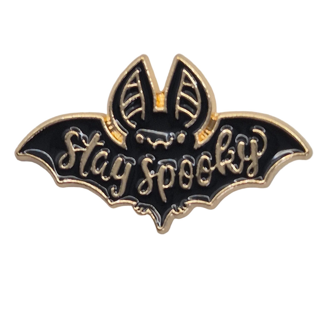 A bat-shaped enamel pin with black enamel and gold-tone metal outlines, featuring the words 'Stay spooky' in raised gold lettering on a white background.