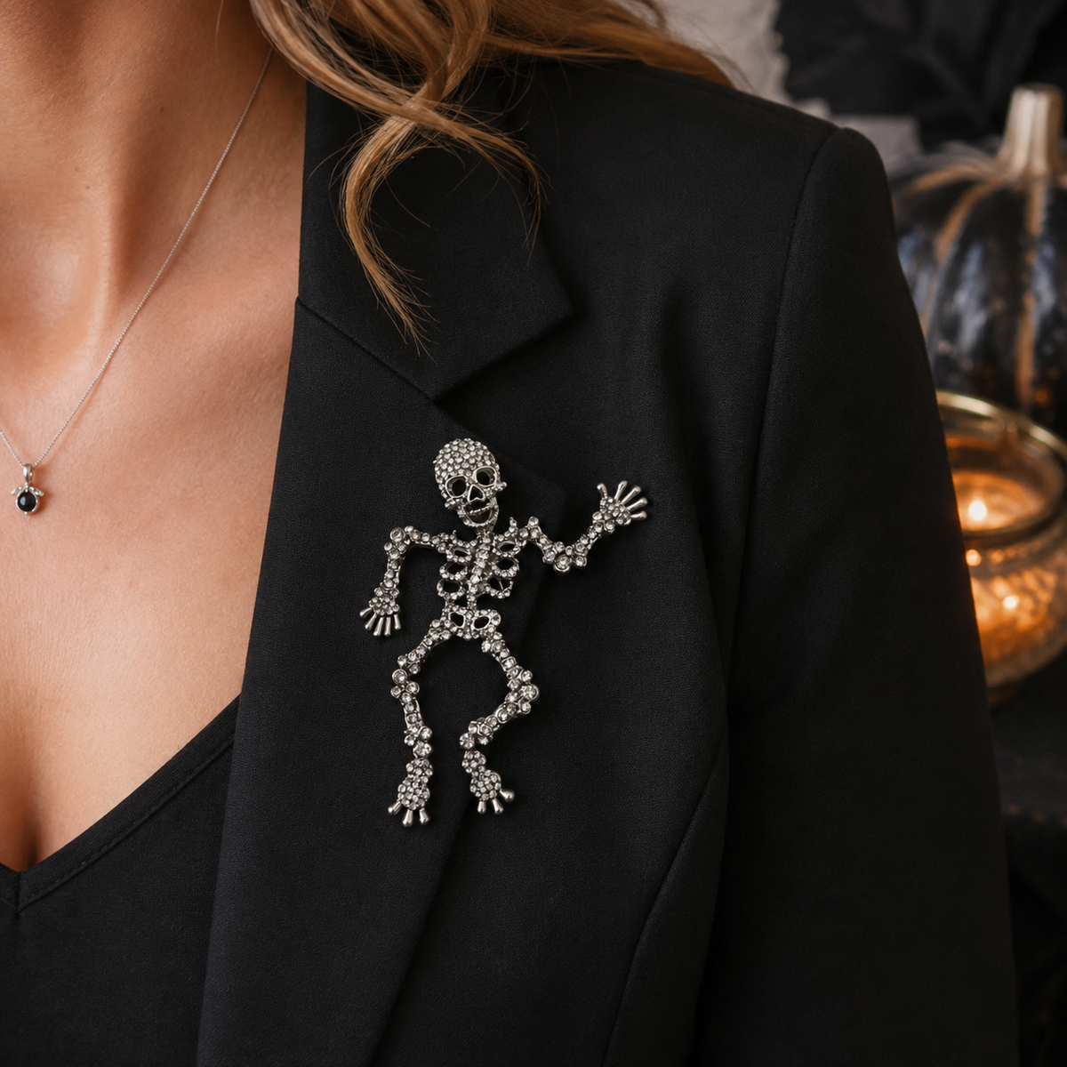 A skeleton-shaped brooch embellished with sparkling clear rhinestones, worn on the lapel of a black blazer. The image is a close-up lifestyle shot showing a model's neck and shoulder, with a warm, dimly lit background featuring a glowing candle and pumpkins.