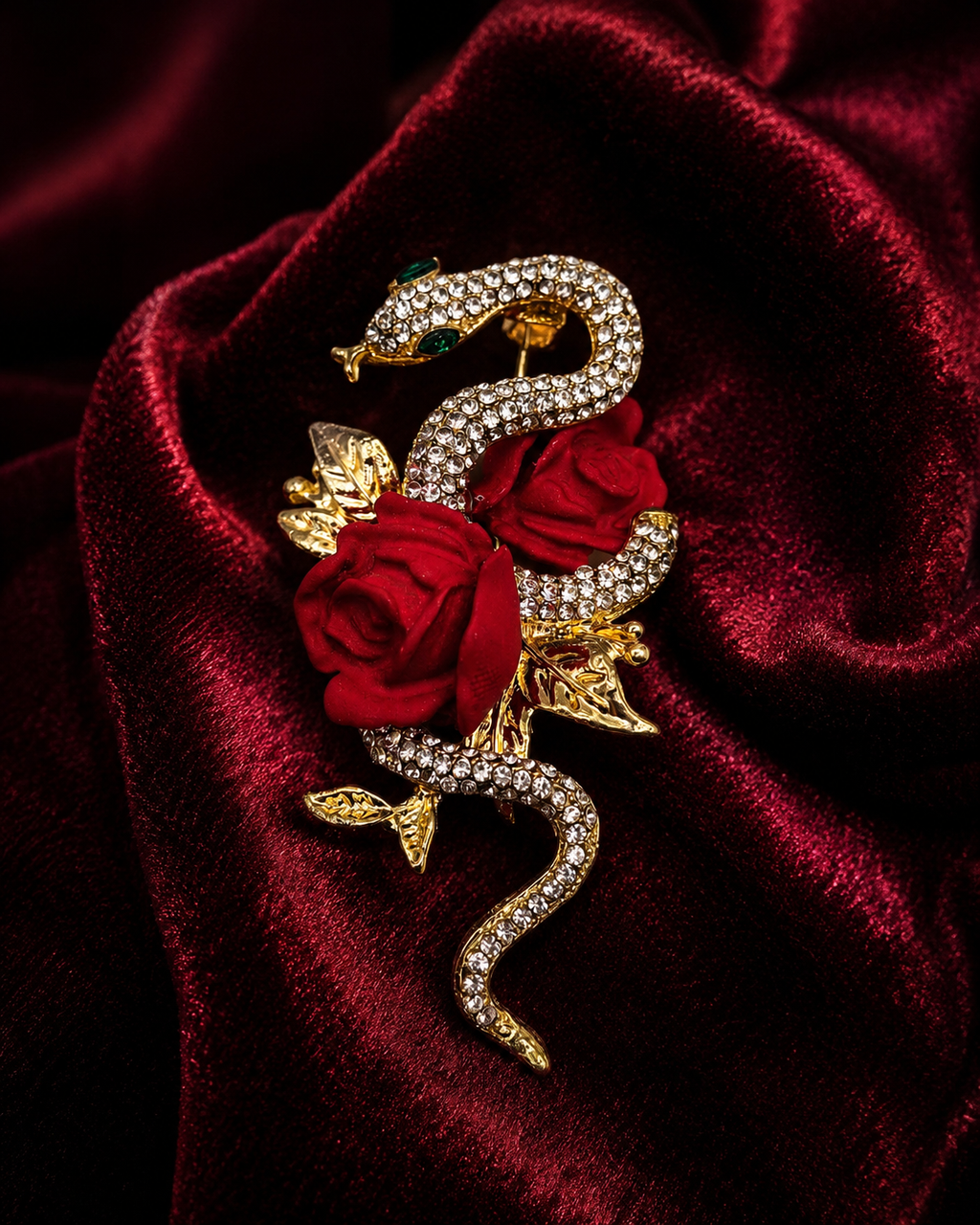 A gold-toned snake brooch embellished with clear rhinestones and accented with two red roses and gold leaves, displayed on a draped dark red velvet fabric.