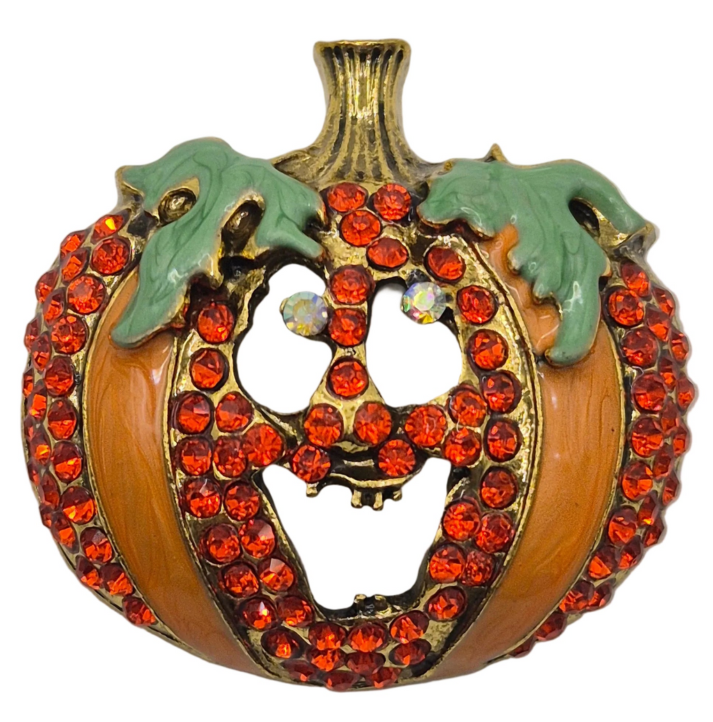 A Halloween pumpkin-shaped brooch or pin with a carved jack-o'-lantern face, covered in orange-red rhinestones, with green enamel leaves and a gold-tone stem on a white background.