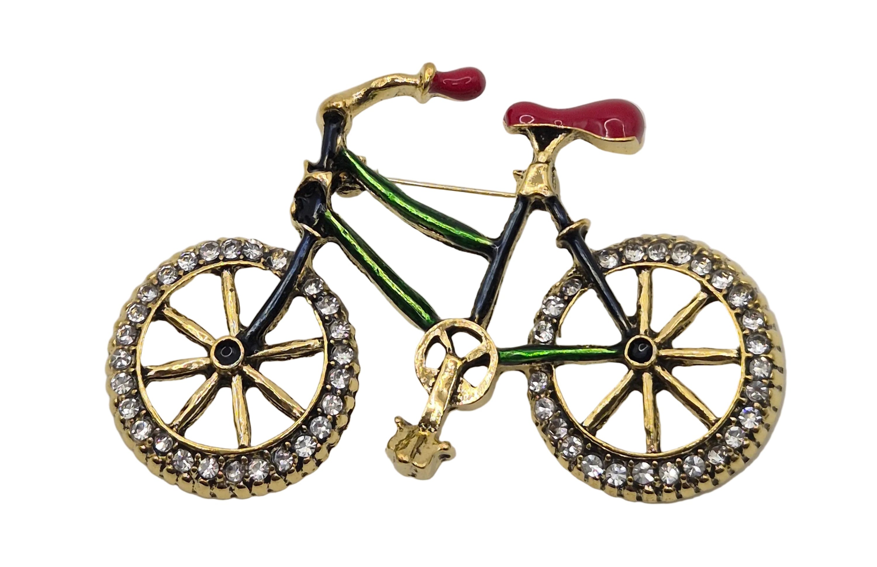 A bicycle-shaped brooch featuring gold-tone metal with black and green enamel details, a red seat and handlebar grip, and clear rhinestones on the wheel rims, shown on a plain white background.