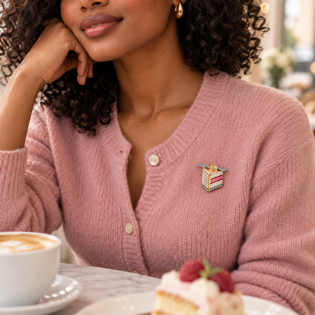 A woman in a cafe wearing a dusty pink knit cardigan with a small colorful slice-of-cake enamel pin on the chest. A cup of coffee and a cake are visible on the table in the foreground.
