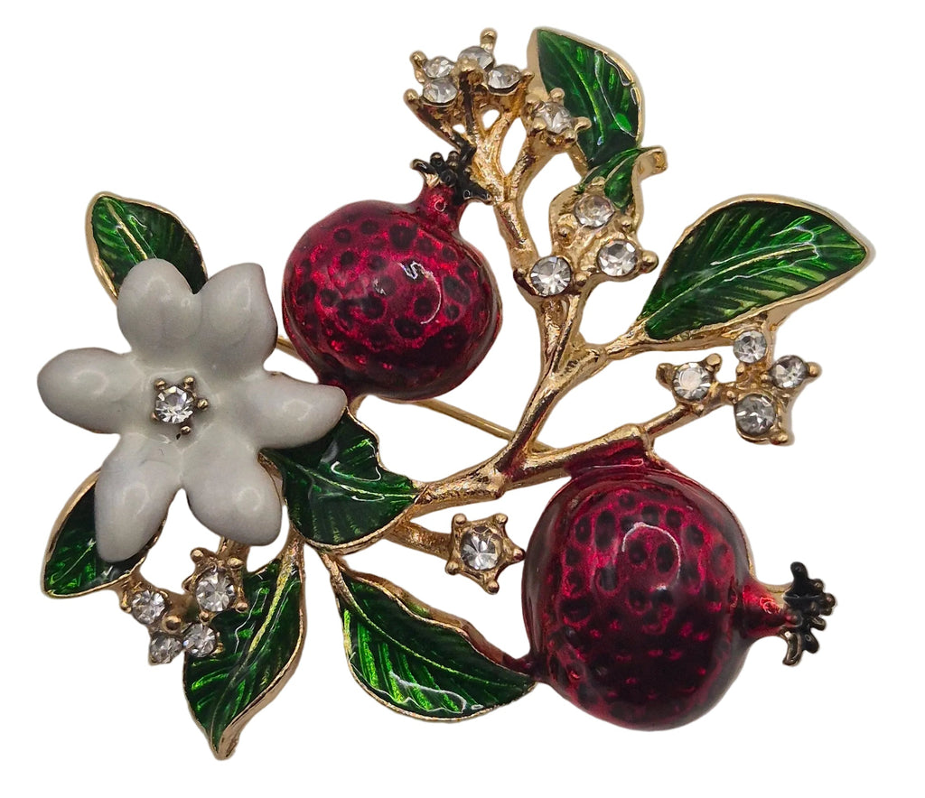 A gold-tone enamel brooch featuring two dark red pomegranate fruits, green leaves, and a white flower with a clear center stone, set against a plain white background.