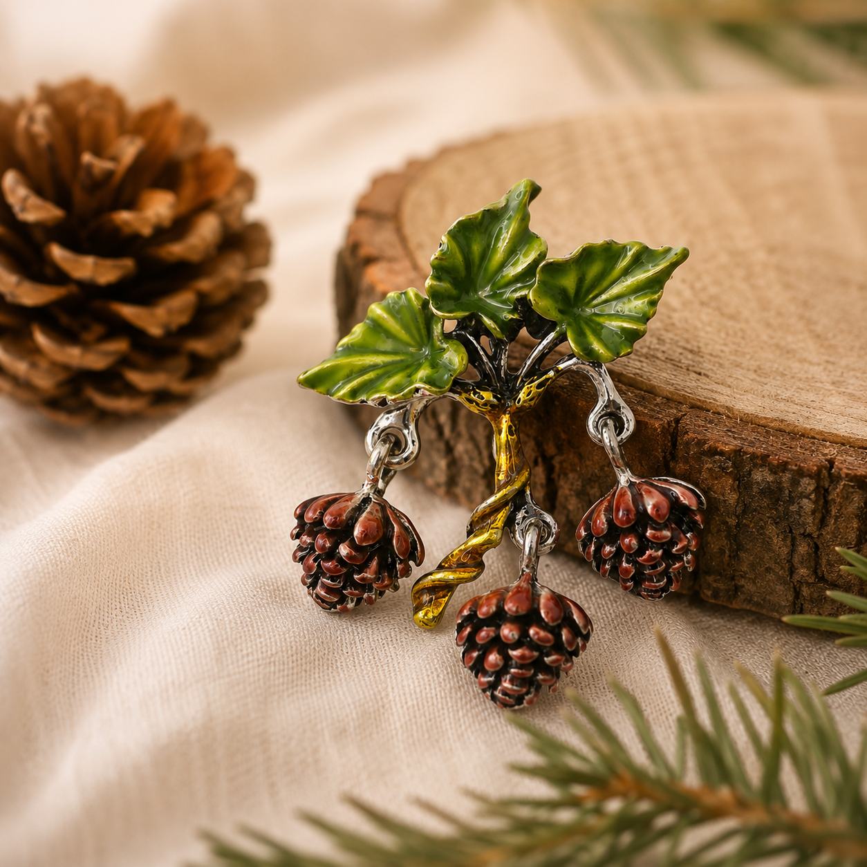 Green enamel pinecone branch brooch with three dangling pinecones and a twisted gold-tone stem, styled on soft cream linen with a rustic wood slice and evergreen accents in a cosy woodland setting.
