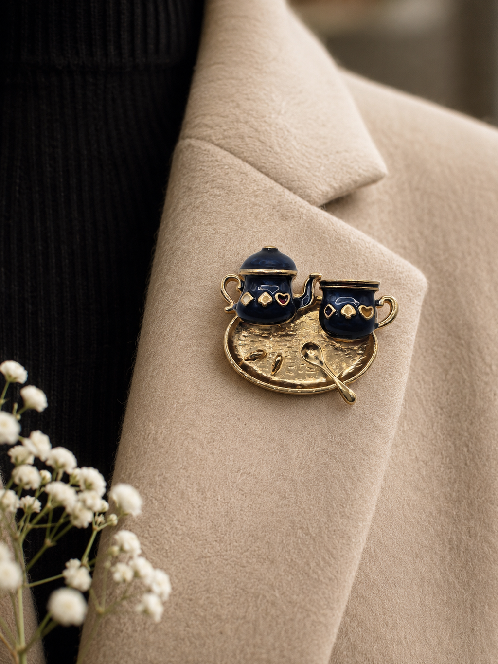A close-up of a gold-toned enamel lapel pin shaped like a coffee pot and cup, pinned onto a beige coat over a black ribbed sweater. Small white flowers appear in the lower left corner.