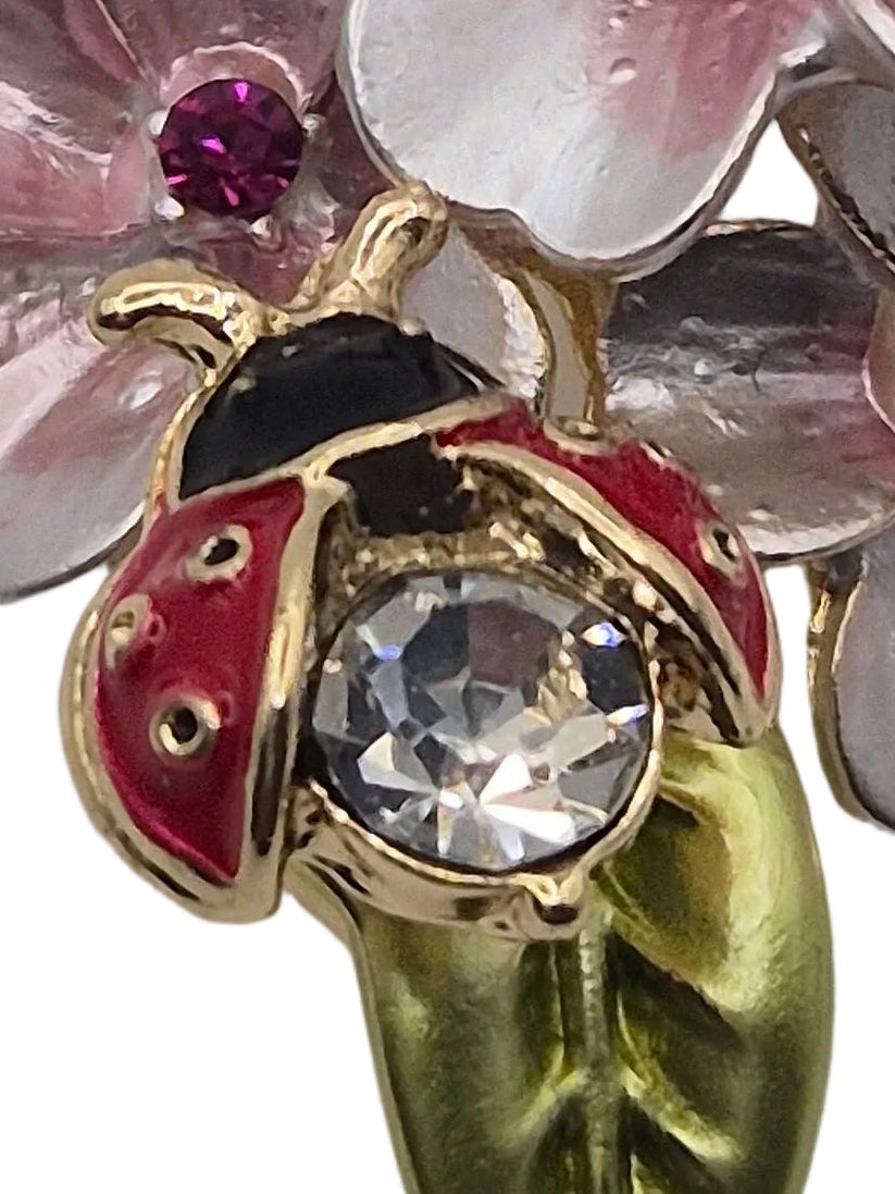 A vintage-style floral brooch featuring three pink and silver cherry blossoms with small red gem accents, a gold leaf stem, and a ladybug detail on a light gray background.