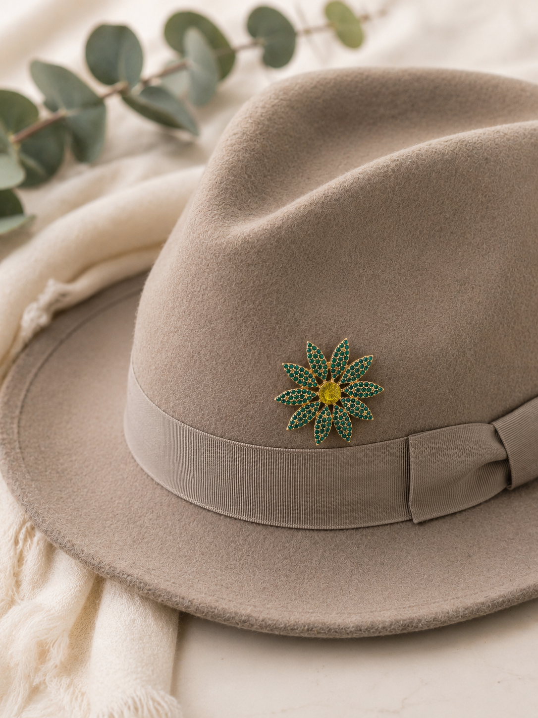 A taupe felt fedora hat with a matching ribbon band and bow, decorated with a green and yellow flower-shaped brooch, styled with a cream scarf and eucalyptus leaves.