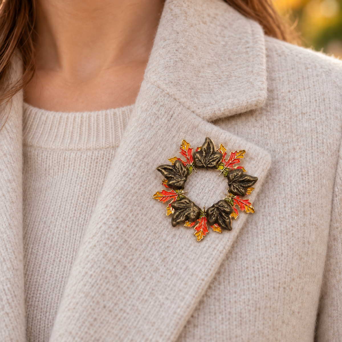 A close-up of a person wearing a circular brooch made of dark metallic leaves with red and gold enamel accents, pinned to the lapel of a cream-colored knit sweater.