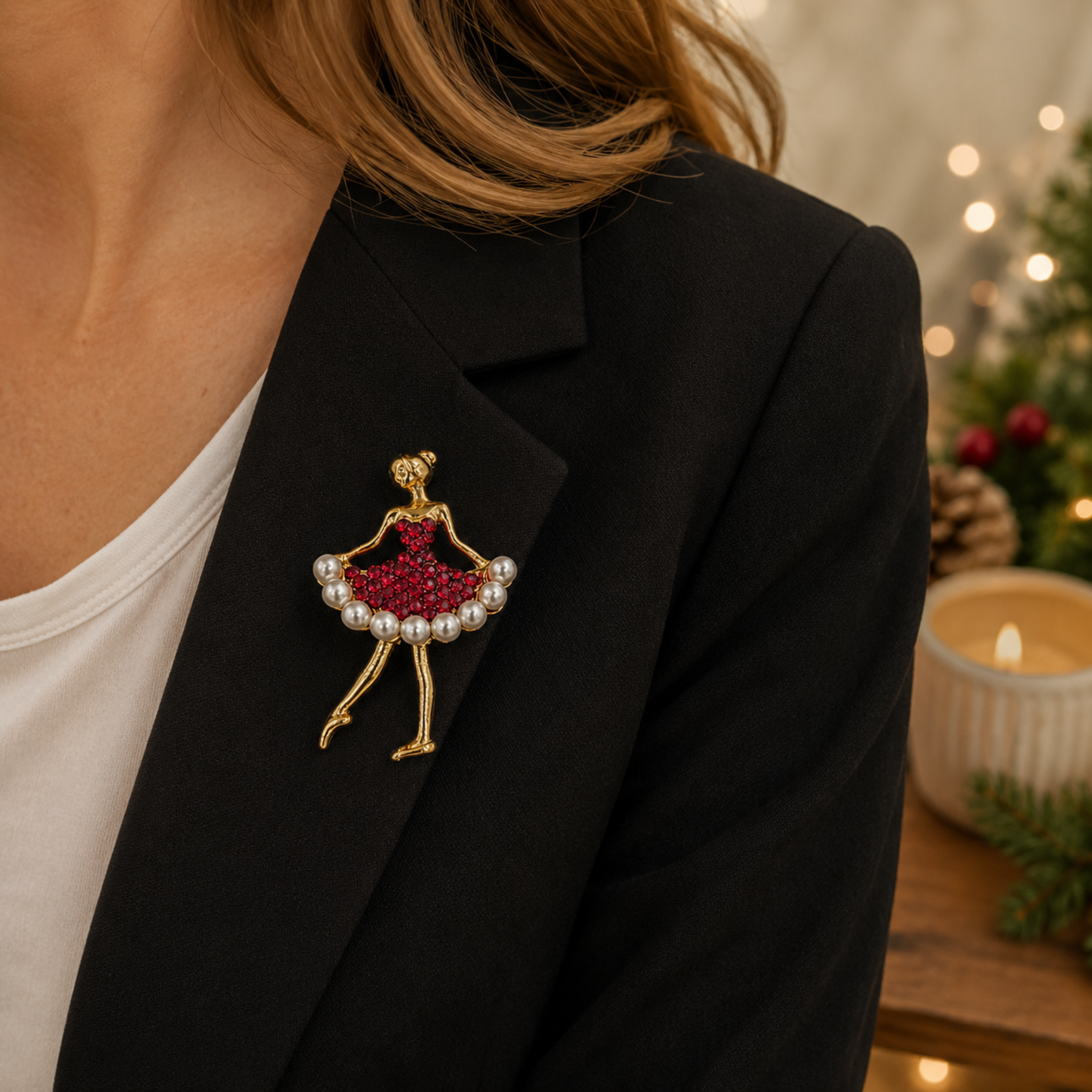 A gold-toned ballerina brooch with a red beaded dress and pearl accents, pinned to a black blazer worn by a model in a cozy, softly lit holiday setting.