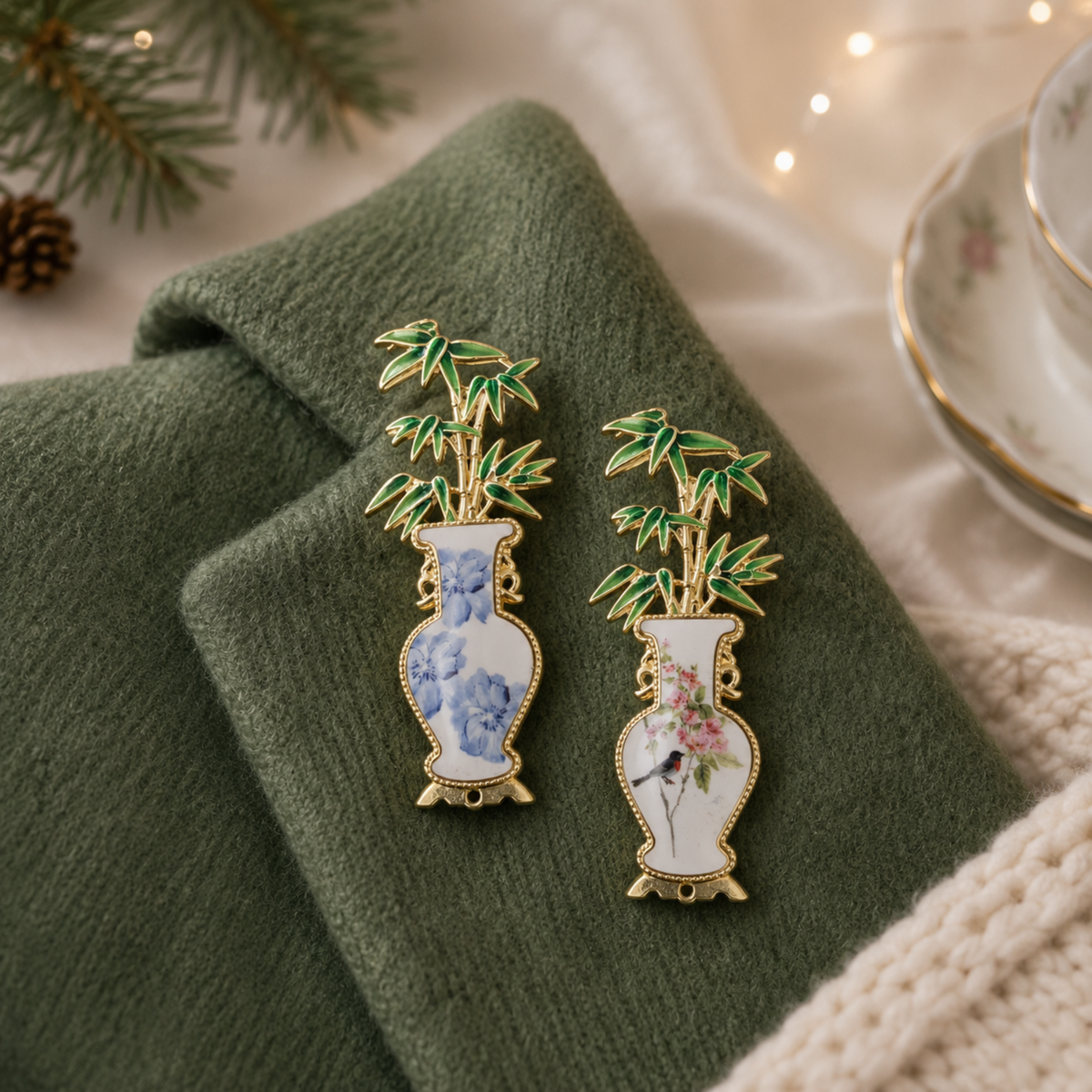 Two enamel lapel pins shaped like white porcelain vases with bamboo foliage, displayed on a folded dark green fabric. One pin features blue floral detailing, and the other features pink floral detailing, both with gold-tone outlines.