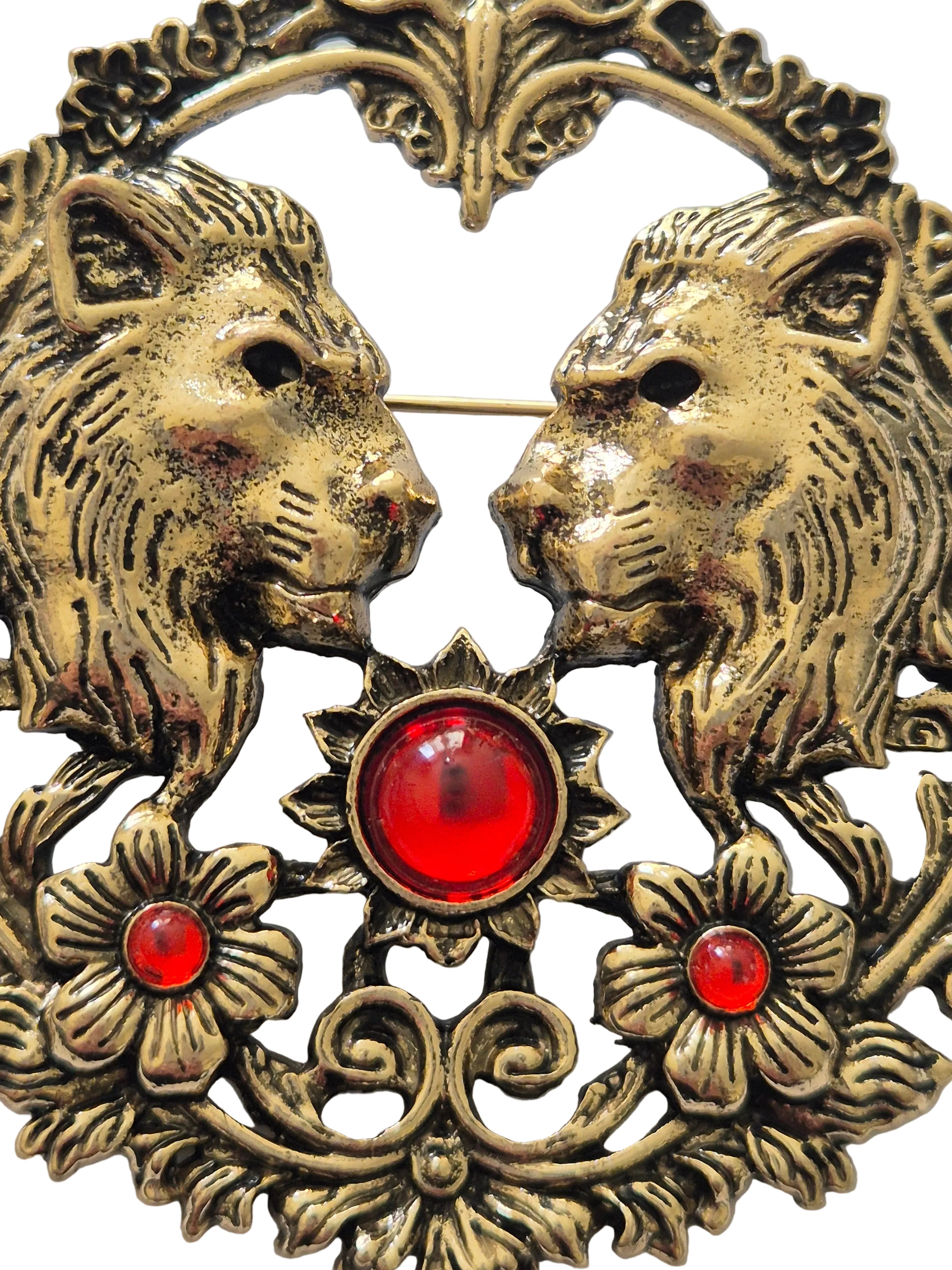 Decorative gold brooch with two lion faces, red gemstones, and floral elements.