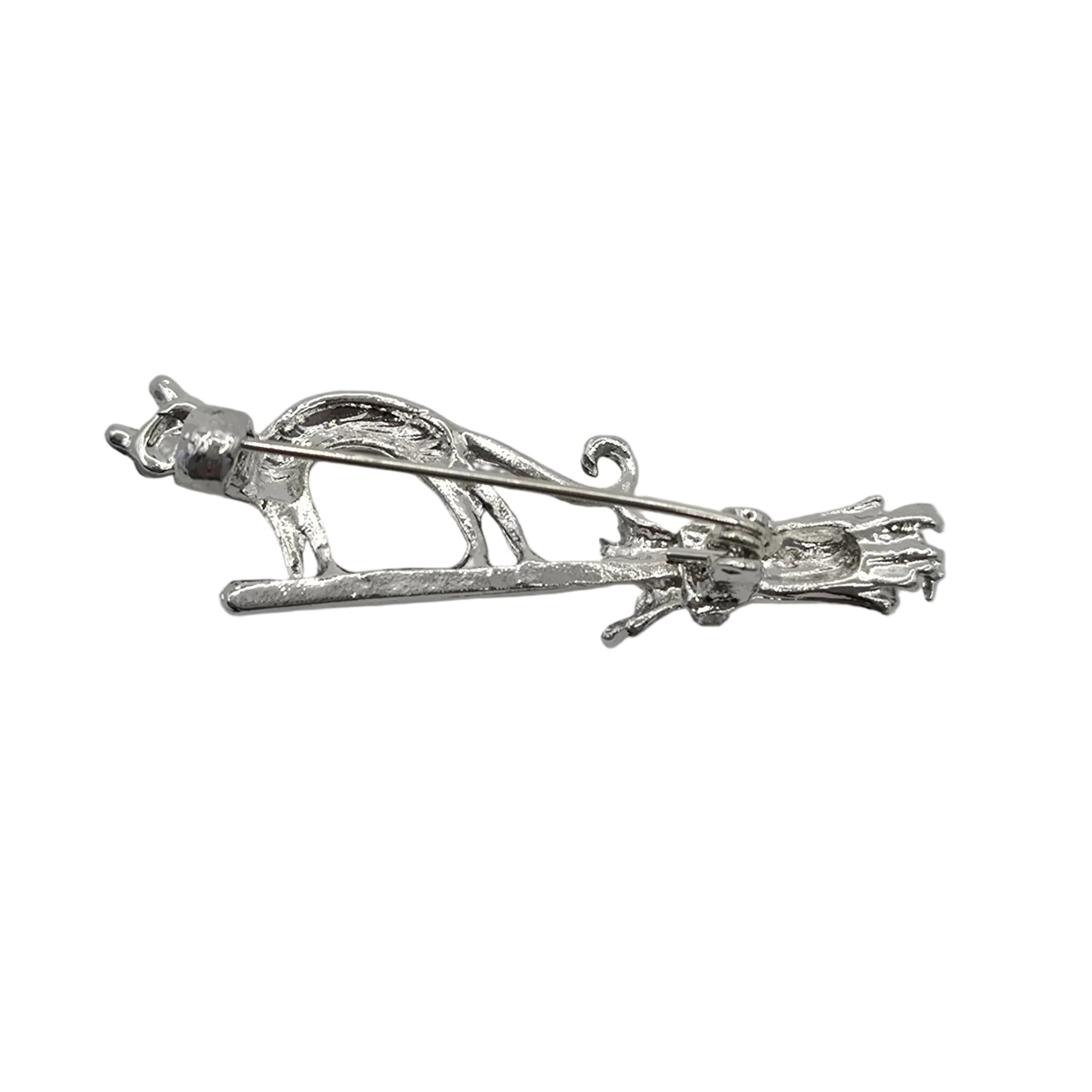 A silver-toned metal brooch featuring a cat standing on a broomstick, displayed against a plain white background.