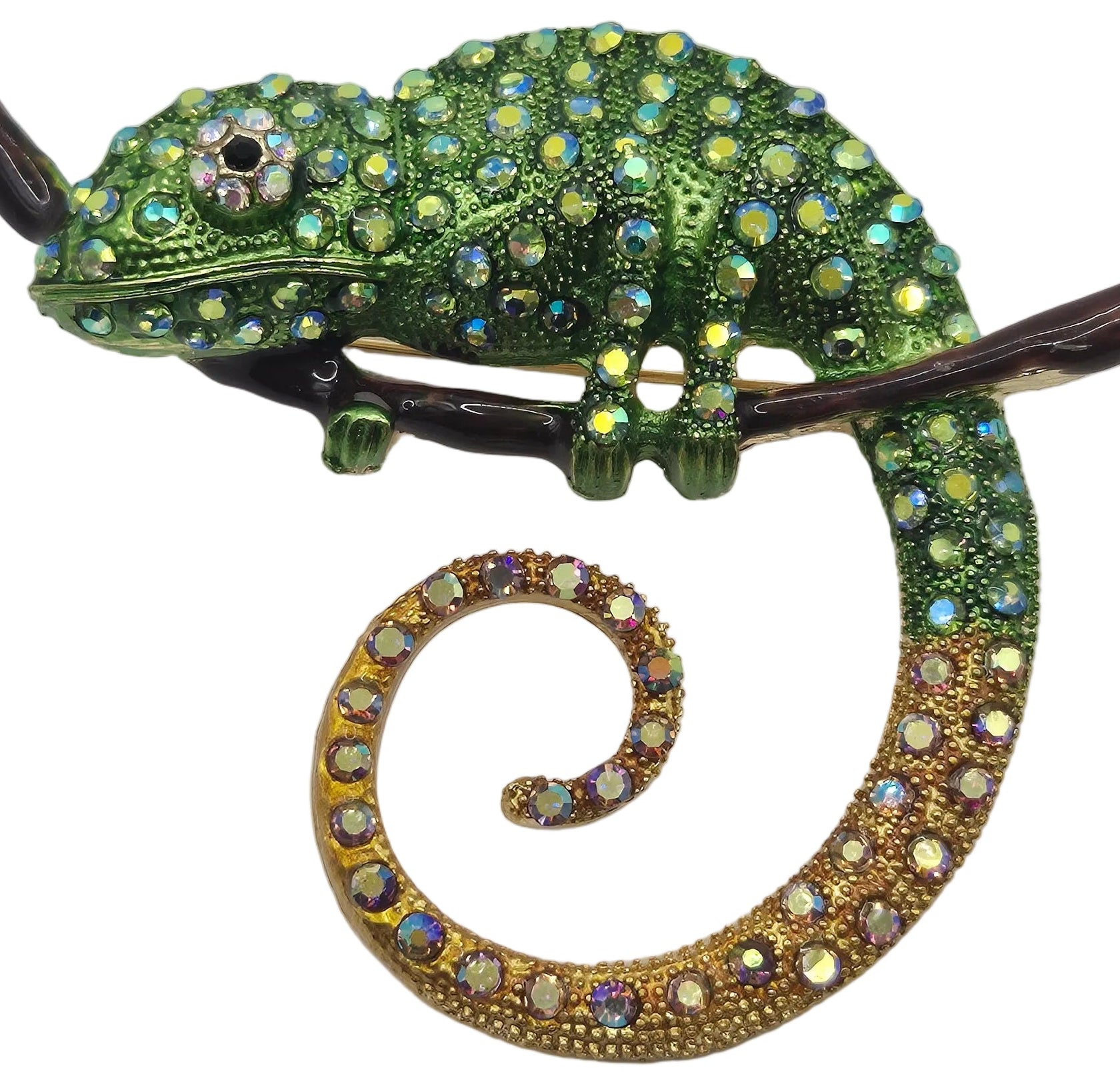 A chameleon-shaped brooch embellished with green rhinestones on its body and head, transitioning to gold rhinestones on its curled tail, perched on a dark brown branch against a white background.