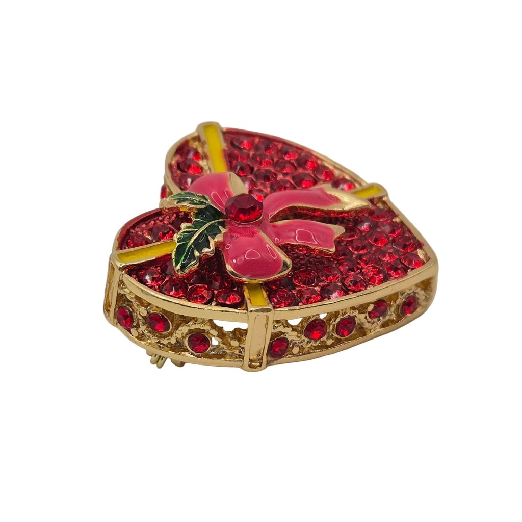 A heart-shaped brooch featuring a gold-tone metal frame, covered in sparkling red rhinestones, with a pink enamel poinsettia flower and green holly leaves on top, accented by yellow enamel details.