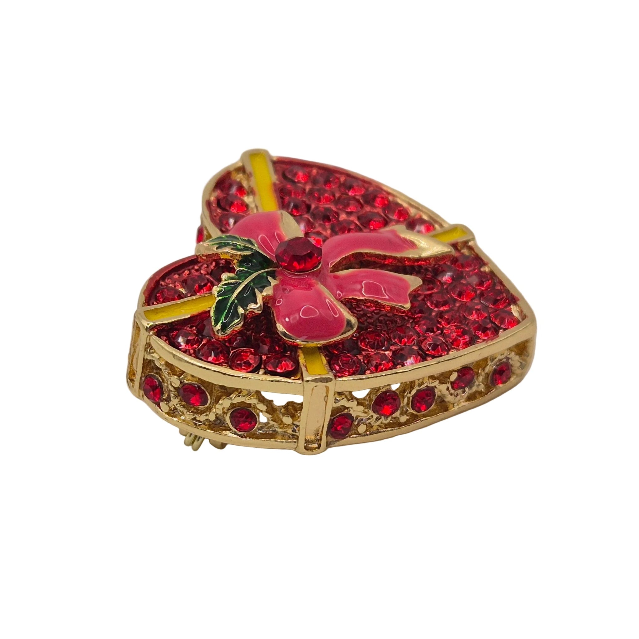 A heart-shaped brooch featuring a gold-tone metal frame, covered in sparkling red rhinestones, with a pink enamel poinsettia flower and green holly leaves on top, accented by yellow enamel details.