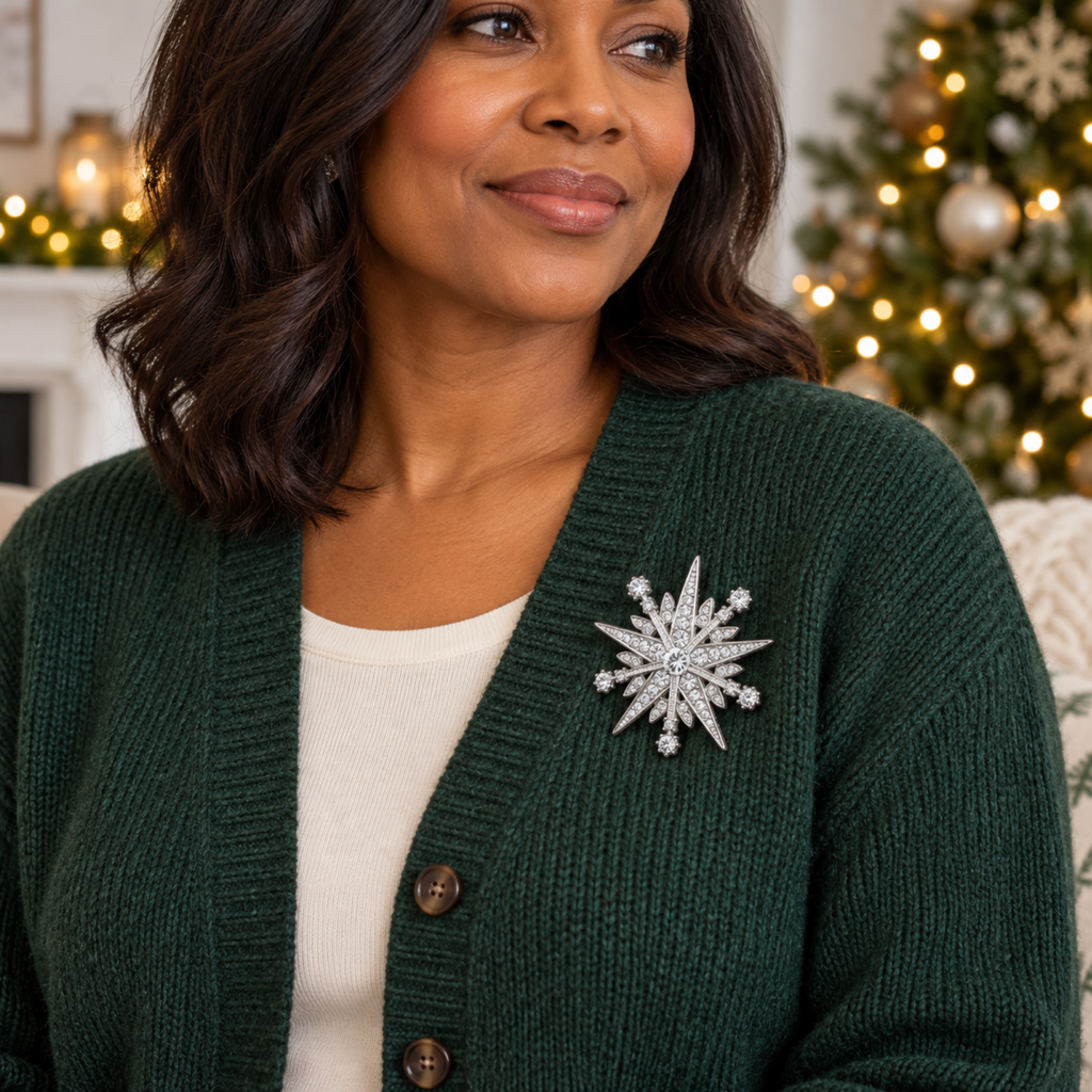 A woman wearing a dark green knit cardigan over a cream top, featuring a sparkling silver snowflake brooch pinned to the chest. The background shows a softly blurred festive holiday scene with a decorated Christmas tree and warm lights.