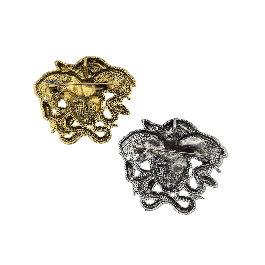 Two medusa head brooches shown on a plain white background, one in gold-tone metal and the other in silver-tone metal.