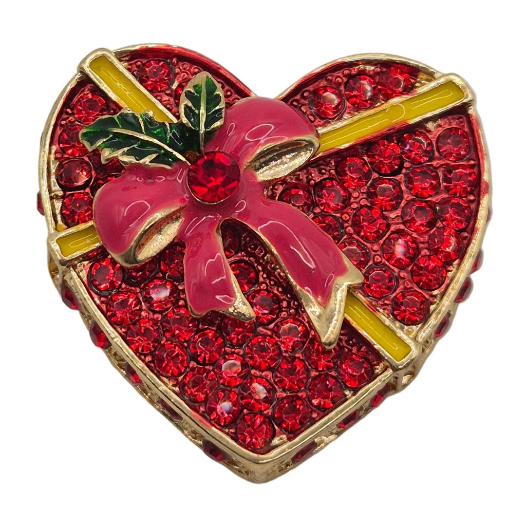 A heart-shaped brooch designed to look like a gift box, covered in red rhinestones with a pink bow, green holly leaves, and gold trim, set against a plain white background.