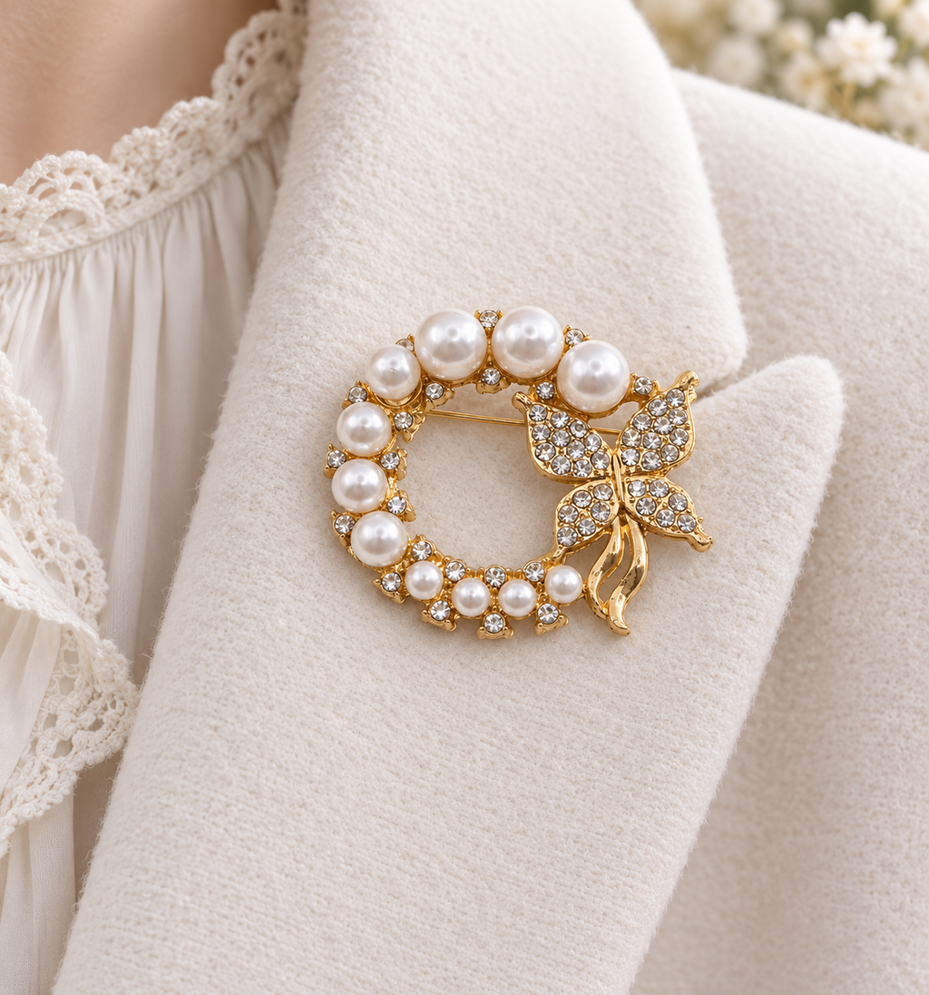 A gold-tone brooch featuring a circular pearl wreath and a butterfly embellished with clear crystals, pinned onto a cream-colored textured jacket.