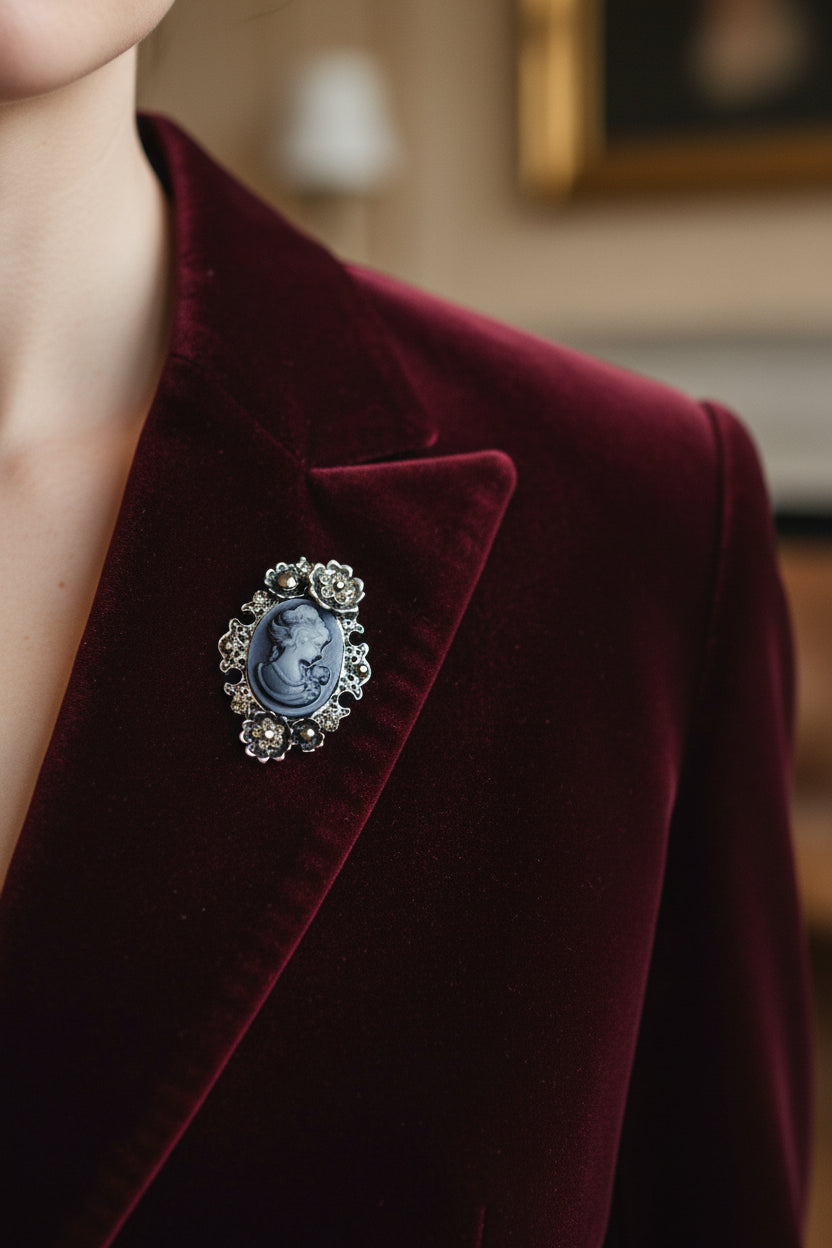 A vintage-style silver brooch featuring a dark blue-gray carved cameo oval of a woman and child, surrounded by floral silver settings with small clear rhinestones, shown on a deep red velvet blazer.