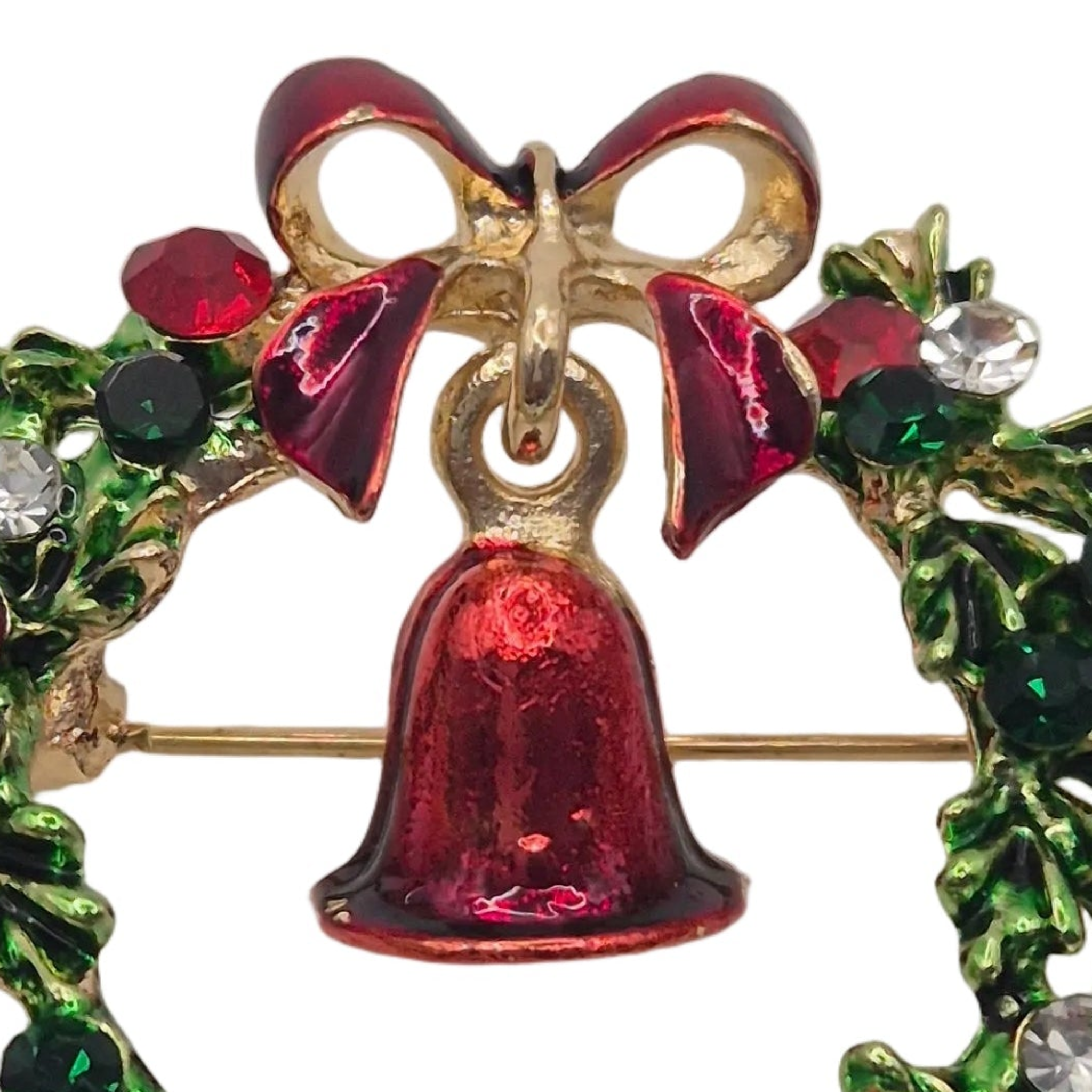 A close-up of a Christmas wreath brooch featuring a red enamel bell, a red bow, green holly leaves, and clear and dark green rhinestones on a white background.