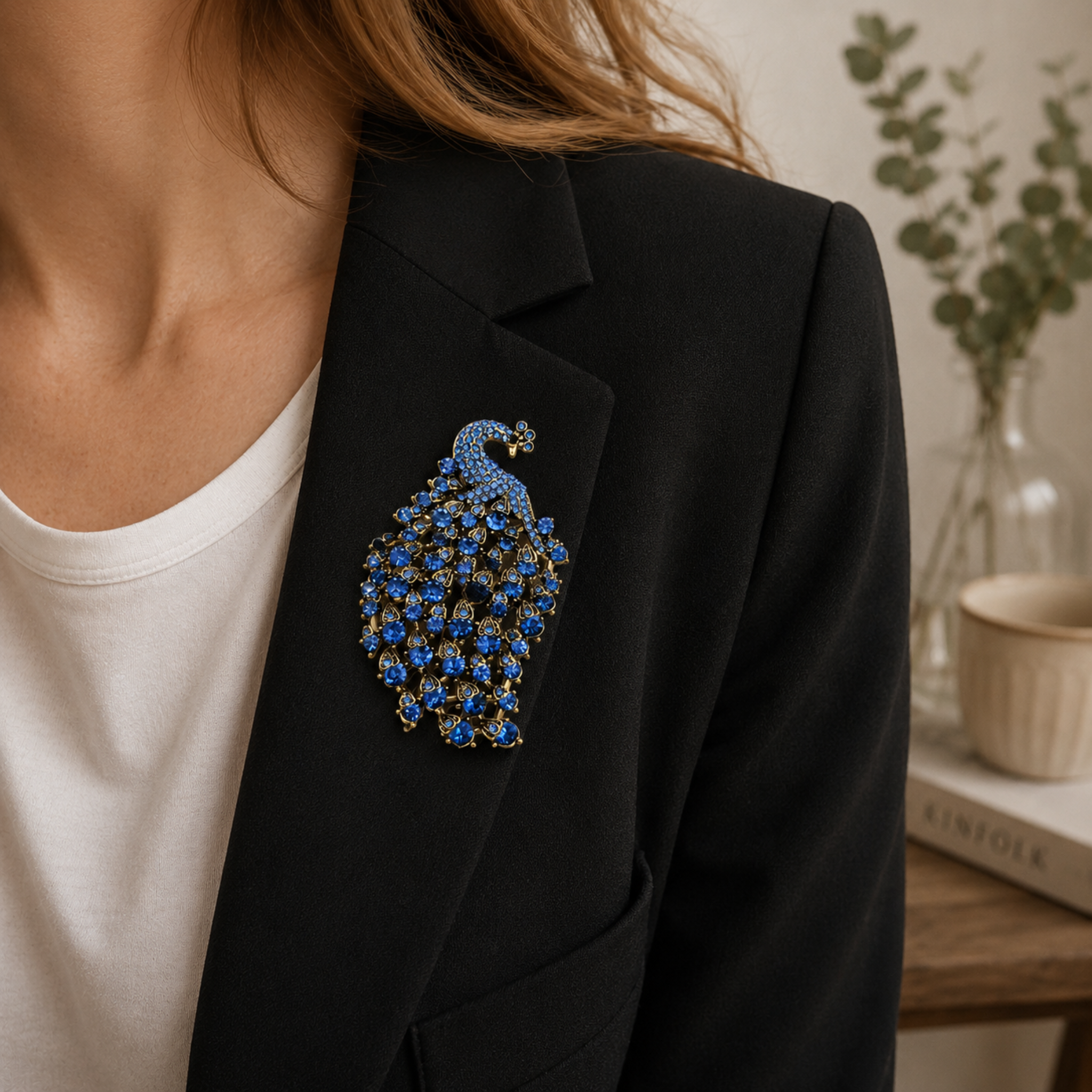 A close-up of a person wearing a black blazer over a white top, featuring a detailed peacock brooch with blue and gold rhinestones on the left lapel.