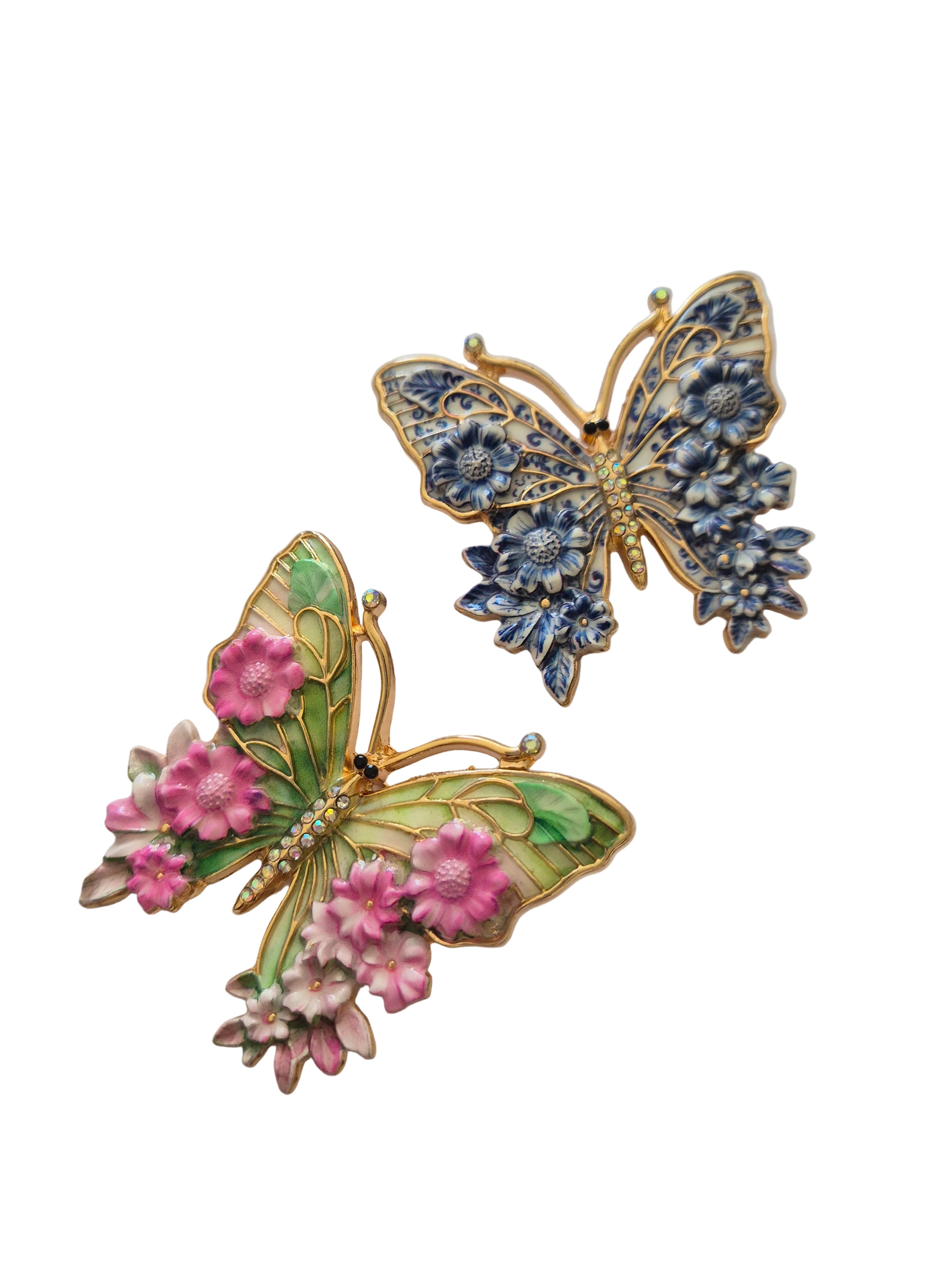 Two butterfly-shaped brooches, one green with pink flowers and the other blue with gold accents, on a white background.